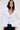 A woman with long dark hair wears the RENAISSANCE RUFFLE DETAIL BLOUSE in white, featuring dramatic ruffles at the neckline and hem, paired with dark denim pants, against a plain light background.
