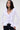 A woman with straight dark hair wears the RENAISSANCE RUFFLE DETAIL BLOUSE, a fitted white top with dramatic ruffles on the deep V-neckline and cuffs, paired with dark jeans against a plain light background.