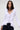 A woman with straight dark hair wears the RENAISSANCE RUFFLE DETAIL BLOUSE—a fitted white top with dramatic ruffles along the neckline and front—paired with dark jeans, standing against a plain light background.