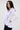 A woman with long dark hair models the RENAISSANCE RUFFLE DETAIL BLOUSE, a fitted white top featuring ruffles along the front and hem, as she stands sideways and looks at the camera against a plain light background.