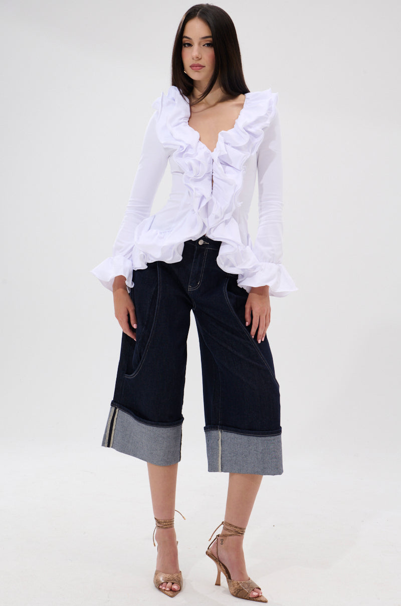 A woman stands against a white background wearing the RENAISSANCE RUFFLE DETAIL BLOUSE with long sleeves and ruffle accents, paired with dark denim wide-leg cuffed capri pants and strappy gold high-heeled sandals.