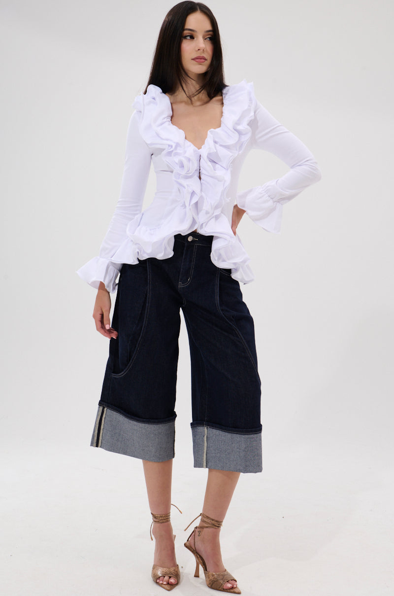 In a studio, a woman models the RENAISSANCE RUFFLE DETAIL BLOUSE in white with long sleeves, paired with wide-leg dark denim culottes, strappy gold heels, and straight dark hair for a romantic look.