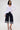 In a studio, a woman models the RENAISSANCE RUFFLE DETAIL BLOUSE in white with long sleeves, paired with wide-leg dark denim culottes, strappy gold heels, and straight dark hair for a romantic look.