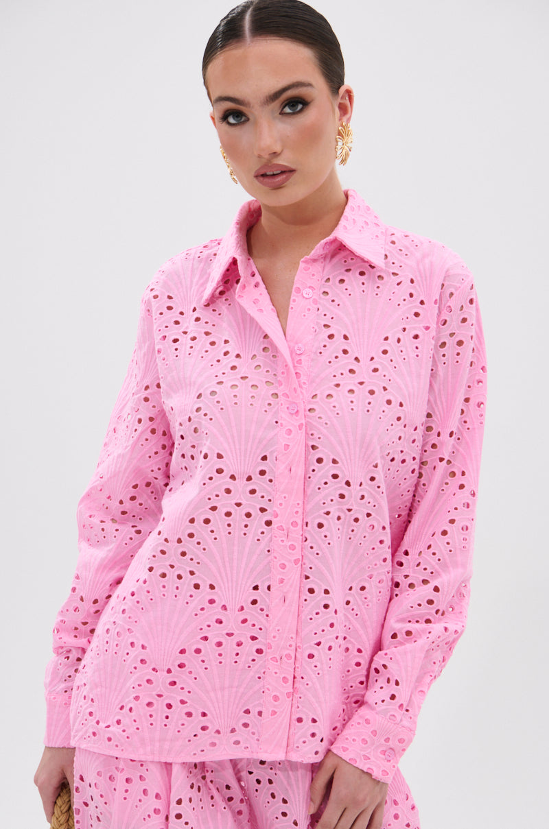 A woman models the JORDAN EYELET BUTTON DOWN TOP in light pink with intricate embroidery, paired with matching bottoms. She wears gold earrings and has slicked-back hair, posing elegantly against a plain white background.