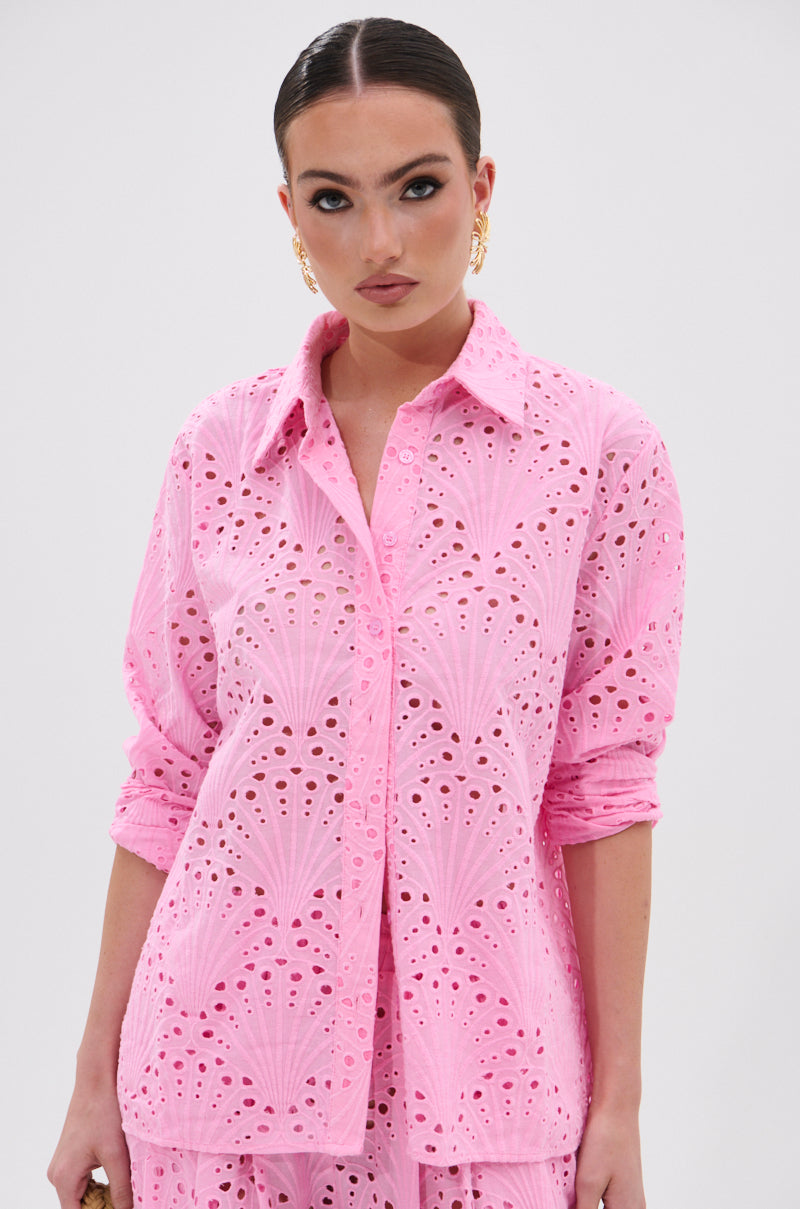 A woman with slicked-back hair and gold hoop earrings wears the JORDAN EYELET BUTTON DOWN TOP, a light pink, long-sleeved shirt with intricate eyelet embroidery, standing against a plain white background.