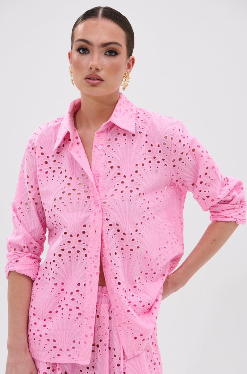 A woman wears the JORDAN EYELET BUTTON DOWN TOP in light pink, featuring long sleeves and a fan embroidery pattern, paired with matching pants as she poses with one hand on her hip against a plain background.