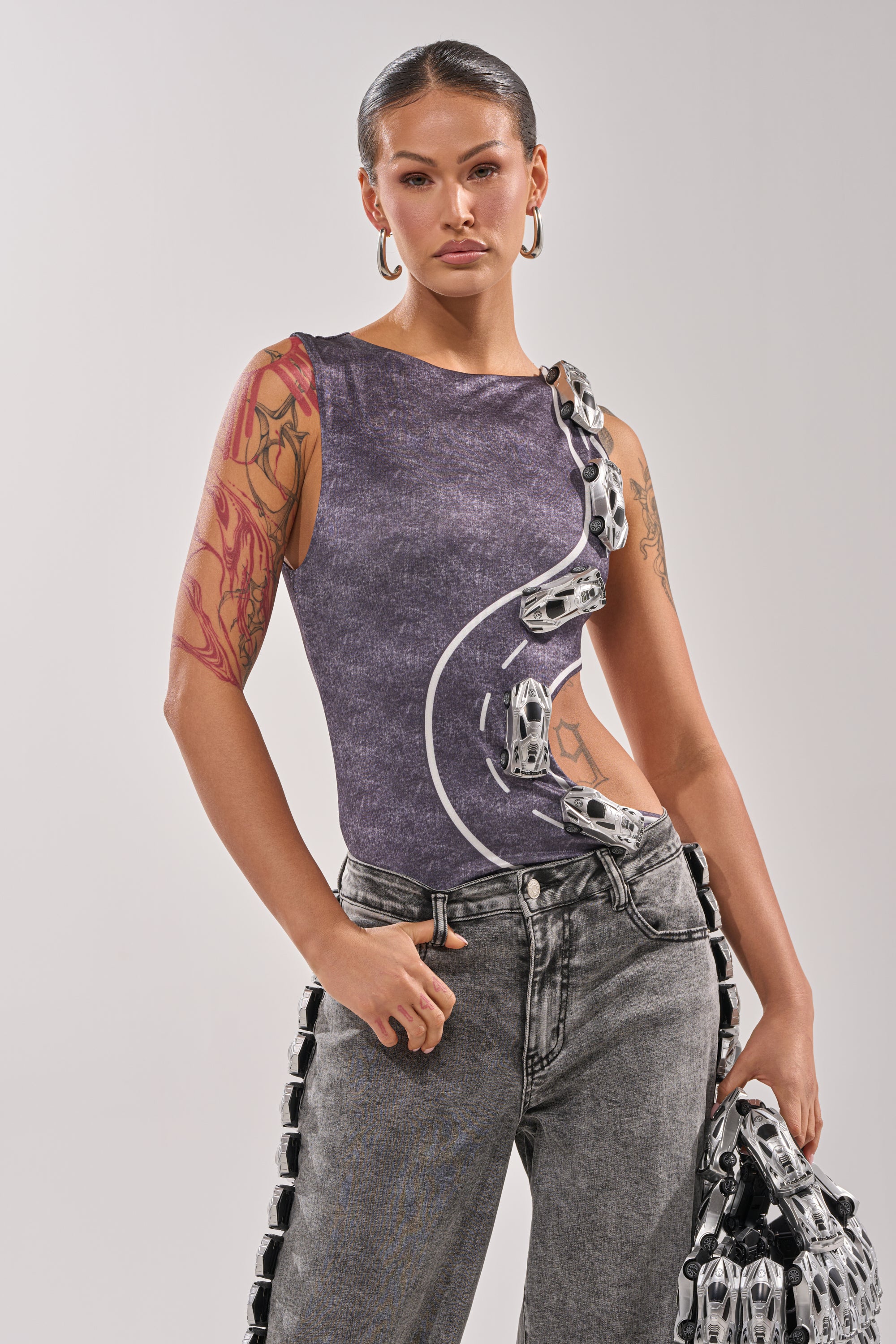 A woman with hoop earrings and tattoos on her left arm wears the OFF TO THE RACES BODYSUIT, a sleeveless purple streetwear piece with metal chain details, paired with high-waisted gray jeans and a matching chain-accented accessory. 0