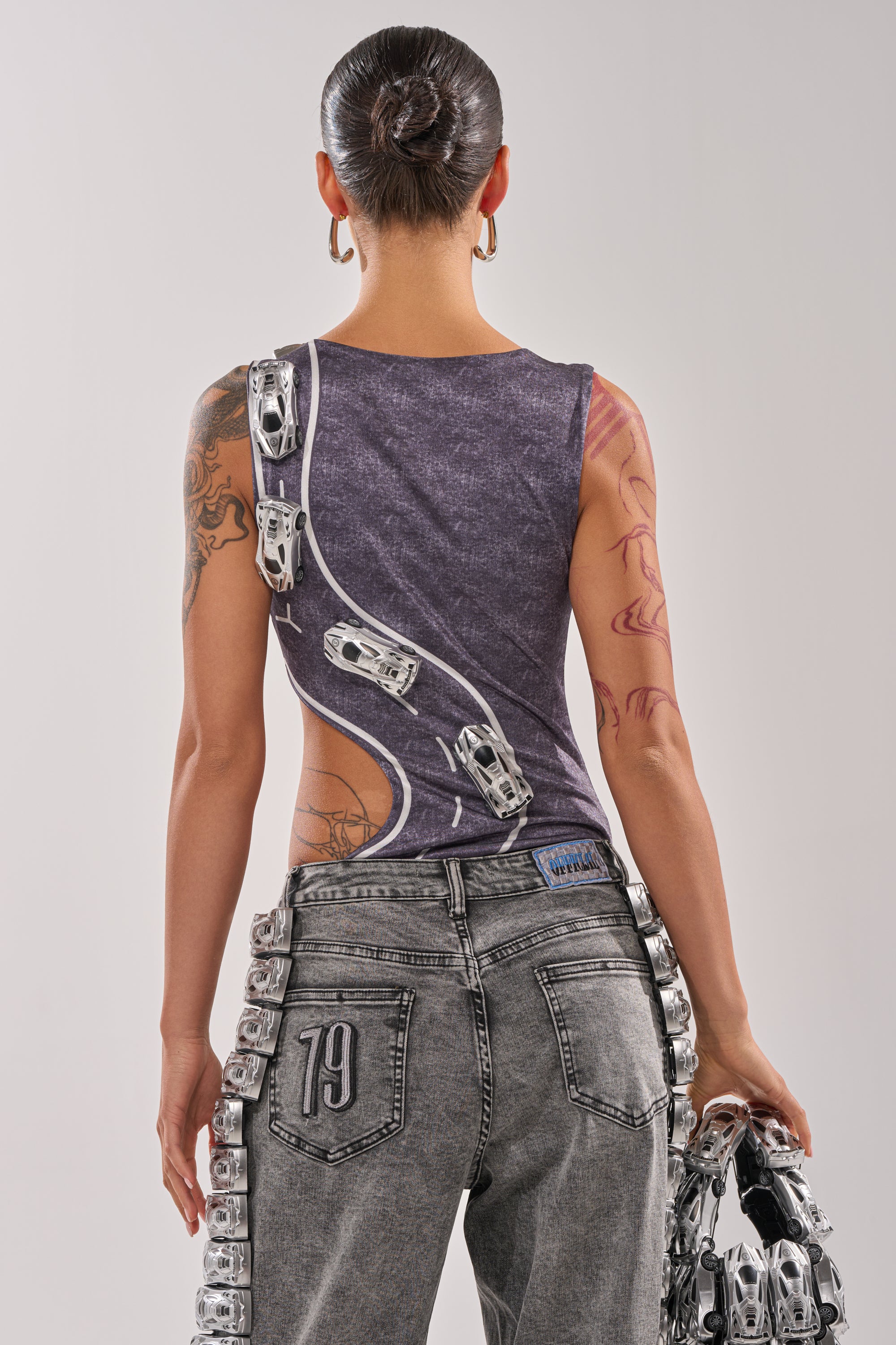 A person faces away wearing the OFF TO THE RACES BODYSUIT, a streetwear-inspired, sleeveless asymmetrical top with metallic buckles, paired with gray jeans marked "79" on the back pocket. They hold a matching metallic accessory and have hair in a bun. 6
