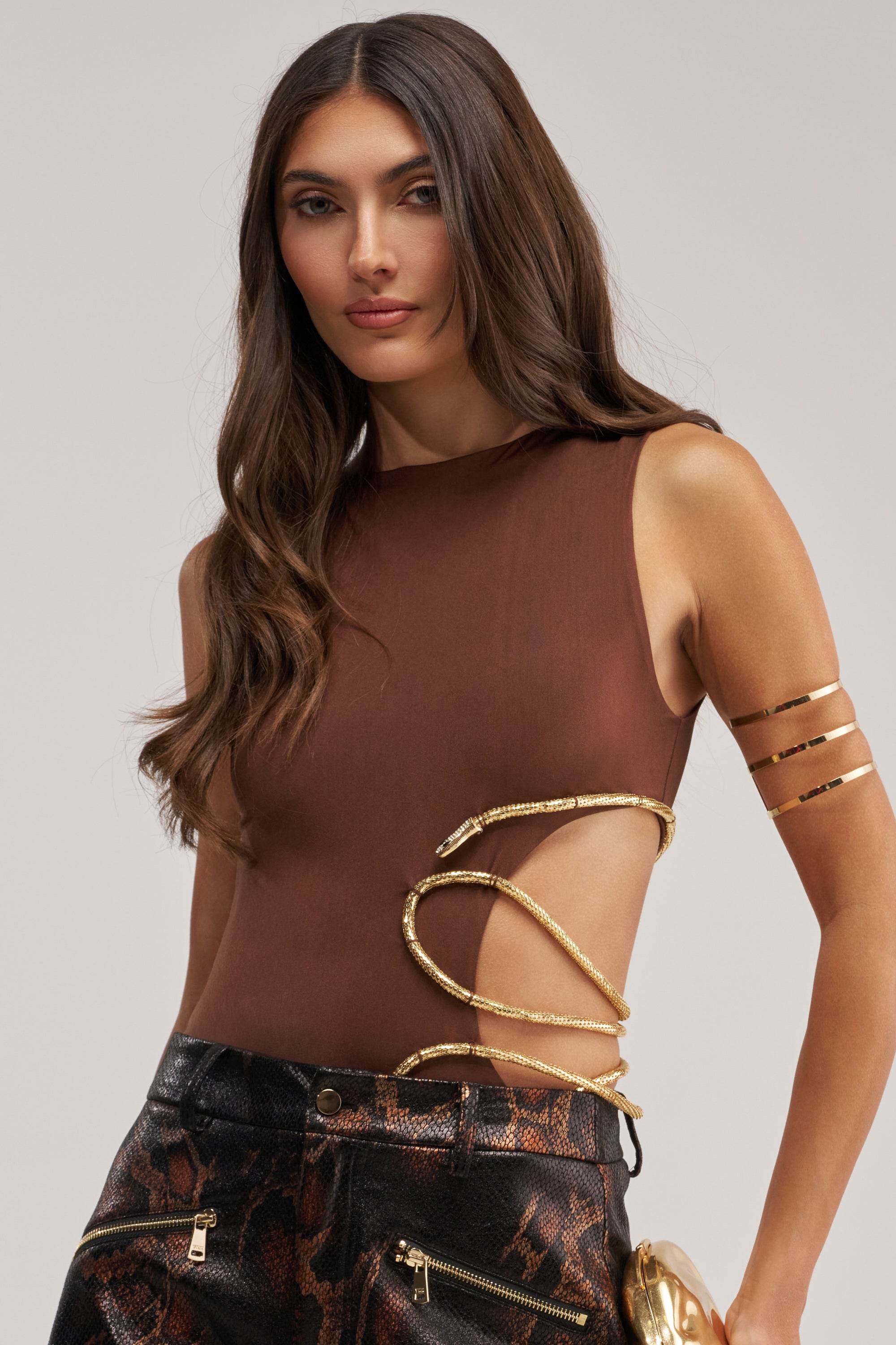 A woman with long brown hair wears the SNEAKY LIL SNAKE BODYSUIT featuring gold accents and pairs it with black snake-print pants. She accessorizes with a gold clutch and arm bands against a light, minimal backdrop—an eye-catching festival look.