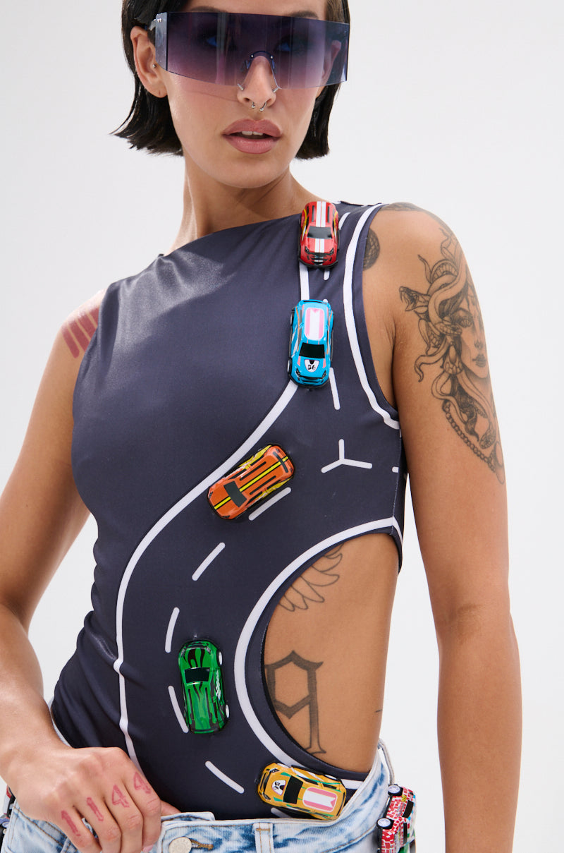 Wearing the FINISH LINE BODYSUIT—a curve-hugging, sleeveless suit with a race track graphic and toy cars—plus oversized sunglasses, nose ring, and arm tattoos, a person poses against a plain background.