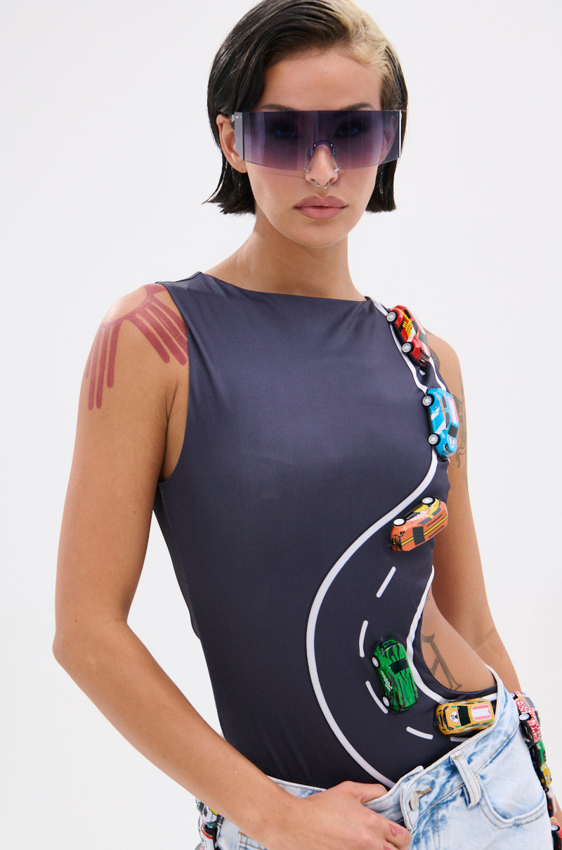 A woman with short dark hair and a blonde streak wears the FINISH LINE BODYSUIT, a curve-hugging black suit with a race track graphic and attached colorful toy cars, paired with large purple sunglasses and jeans.