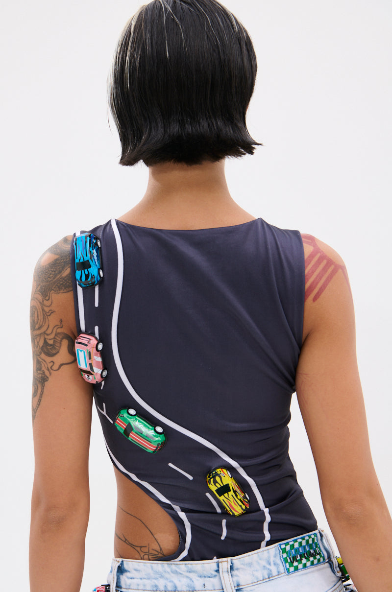 A person with short black and blonde hair faces away, wearing the FINISH LINE BODYSUIT—a curve-hugging suit with a race track and colorful cars graphic. Tattoos are visible on their right arm and shoulder.