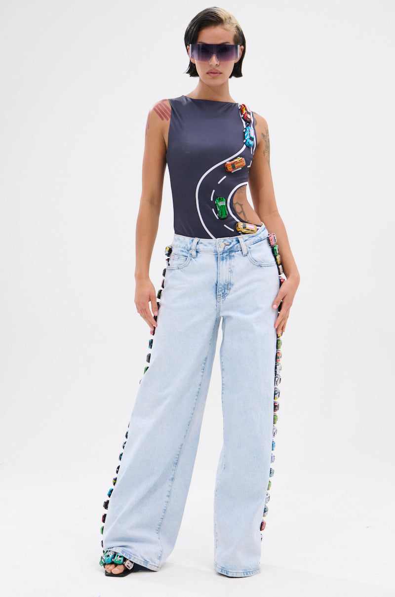 A woman stands confidently against a plain white background in the FINISH LINE BODYSUIT and baggy light blue jeans with toy cars along the seams, her large sunglasses adding bold flair to her look.