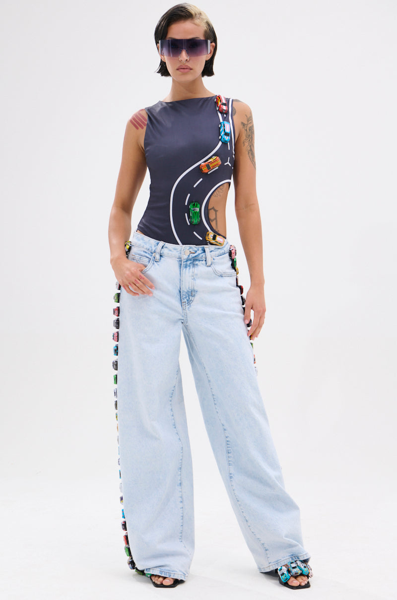 A woman in wide light blue jeans and a curve-hugging FINISH LINE BODYSUIT printed with a race track and toy cars, with toy cars on her jeans and sandals. She wears large sunglasses and has short dark hair.
