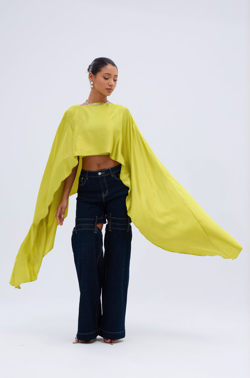 A woman with her hair tied back stands against a plain background, wearing the COCKTAIL HOUR BLOUSE IN CHARTREUSE with an asymmetrical cape design, paired with dark wide-leg jeans featuring white stitching, looking down.