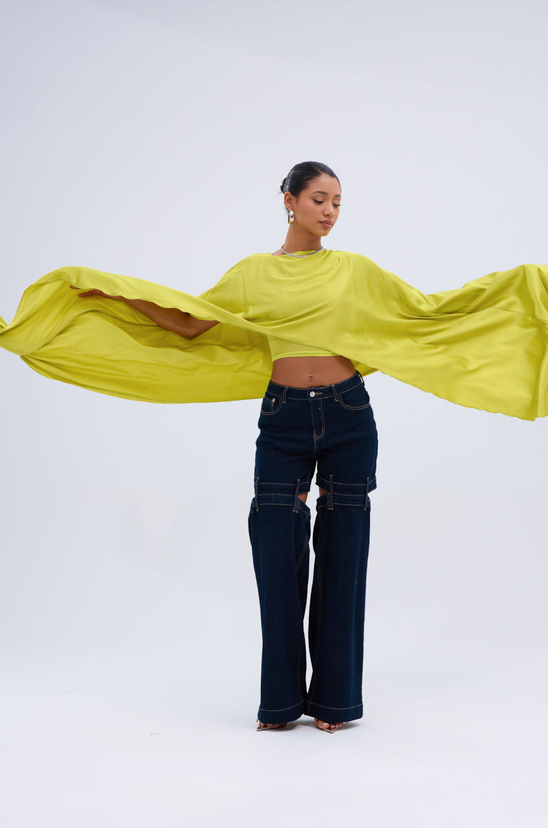 A woman models the COCKTAIL HOUR BLOUSE IN CHARTREUSE, a vibrant yellow-green flowing top with oversized sleeves, paired with dark wide-leg jeans featuring knee cutouts, set against a simple light backdrop.