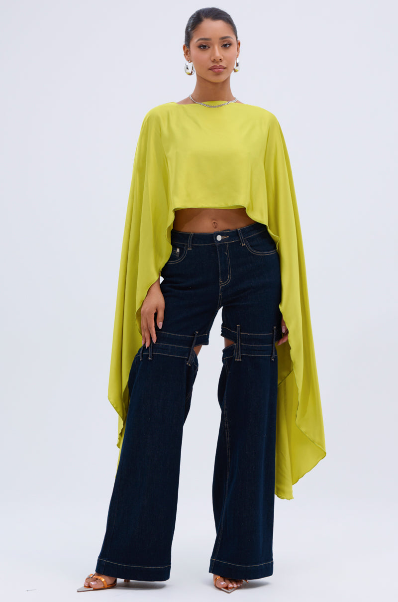 A woman poses in the COCKTAIL HOUR BLOUSE IN CHARTREUSE with flowy long sleeves, paired with high-waisted, wide-leg dark denim jeans featuring cutout and strap details. She completes the look with hoop earrings and sandals against a light backdrop.