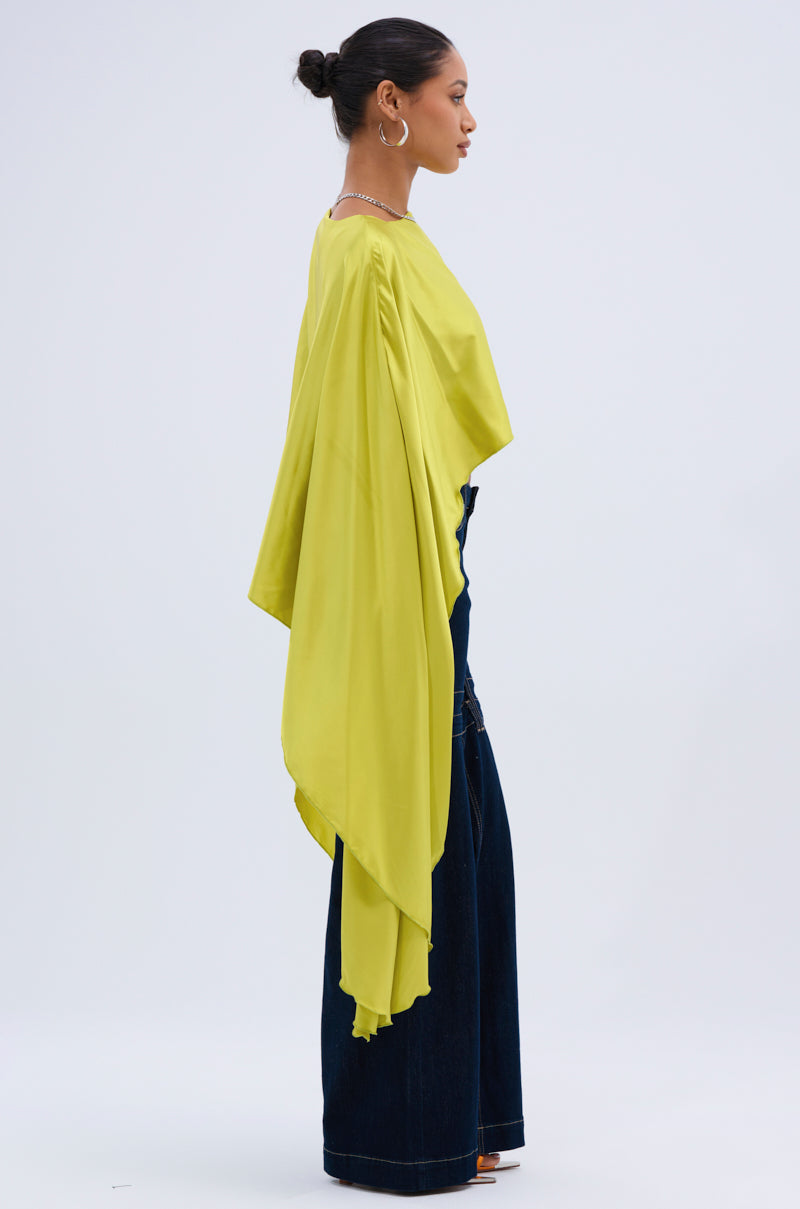 A woman stands in profile wearing the COCKTAIL HOUR BLOUSE IN CHARTREUSE, a satin blouse with a loose back, paired with dark wide-leg jeans and brown open-toe heels. Her hair is in a bun and she wears hoop earrings.