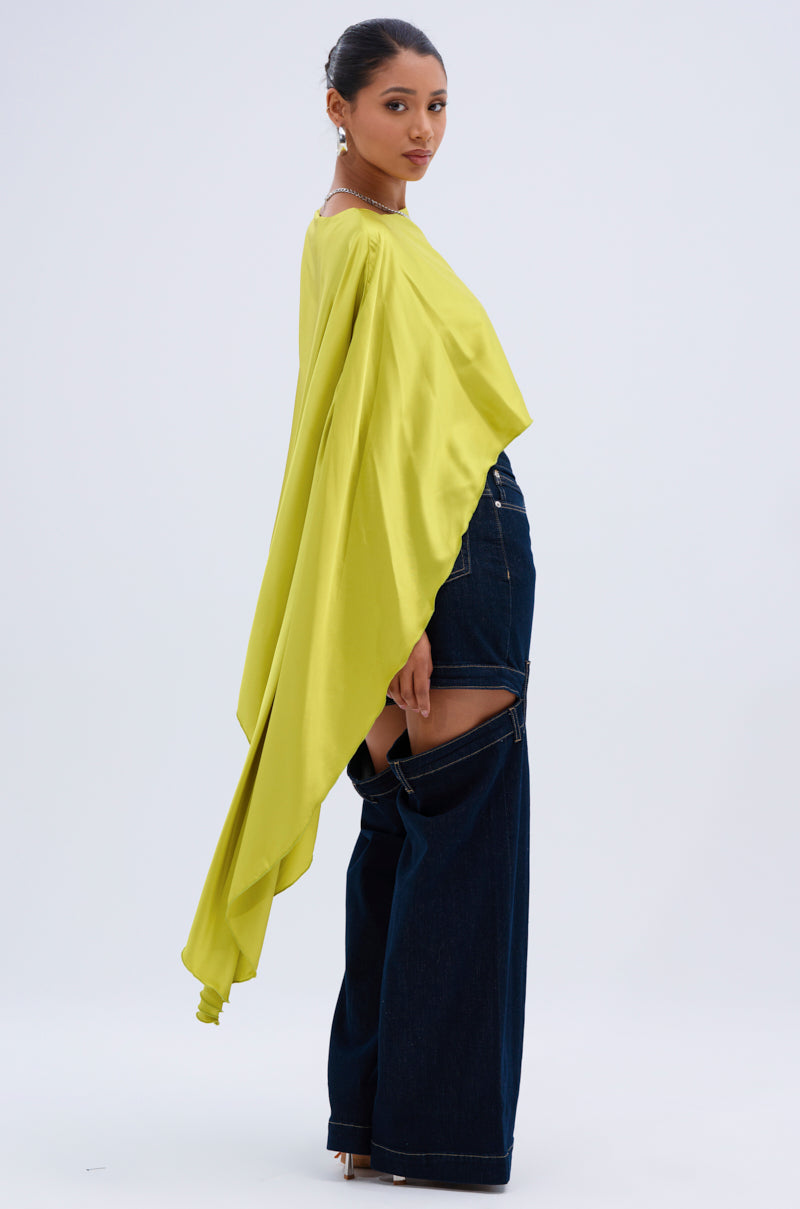 A woman poses in the COCKTAIL HOUR BLOUSE IN CHARTREUSE, a satin top with dramatic asymmetrical draping, styled with wide-leg dark blue jeans featuring large hip cutouts. She stands sideways against a plain light background.