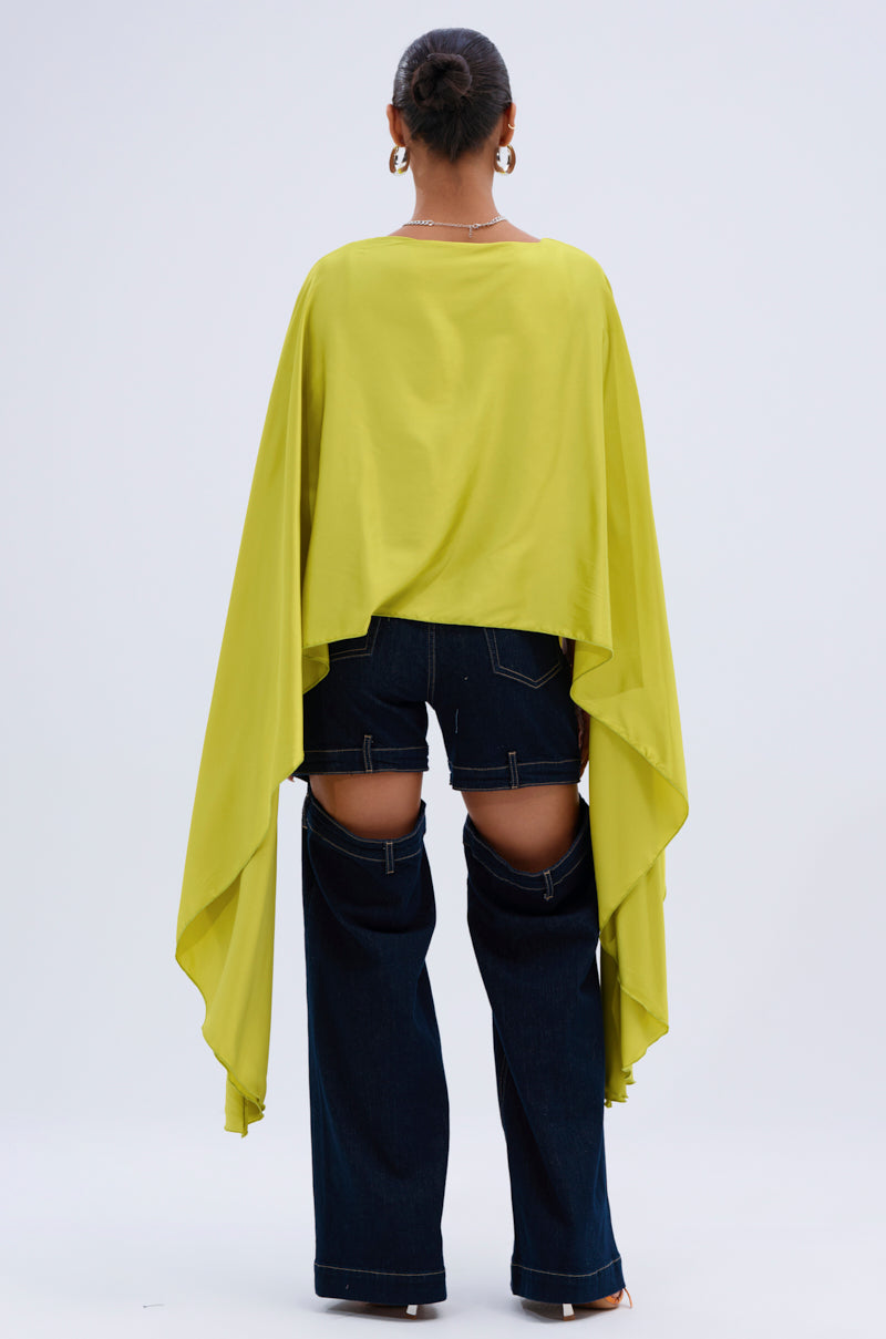 A person stands facing away, wearing the COCKTAIL HOUR BLOUSE IN CHARTREUSE with long draped sleeves and dark blue open-panel jeans that reveal their thighs. Their hair is in a bun, with earrings and heeled shoes completing the look.