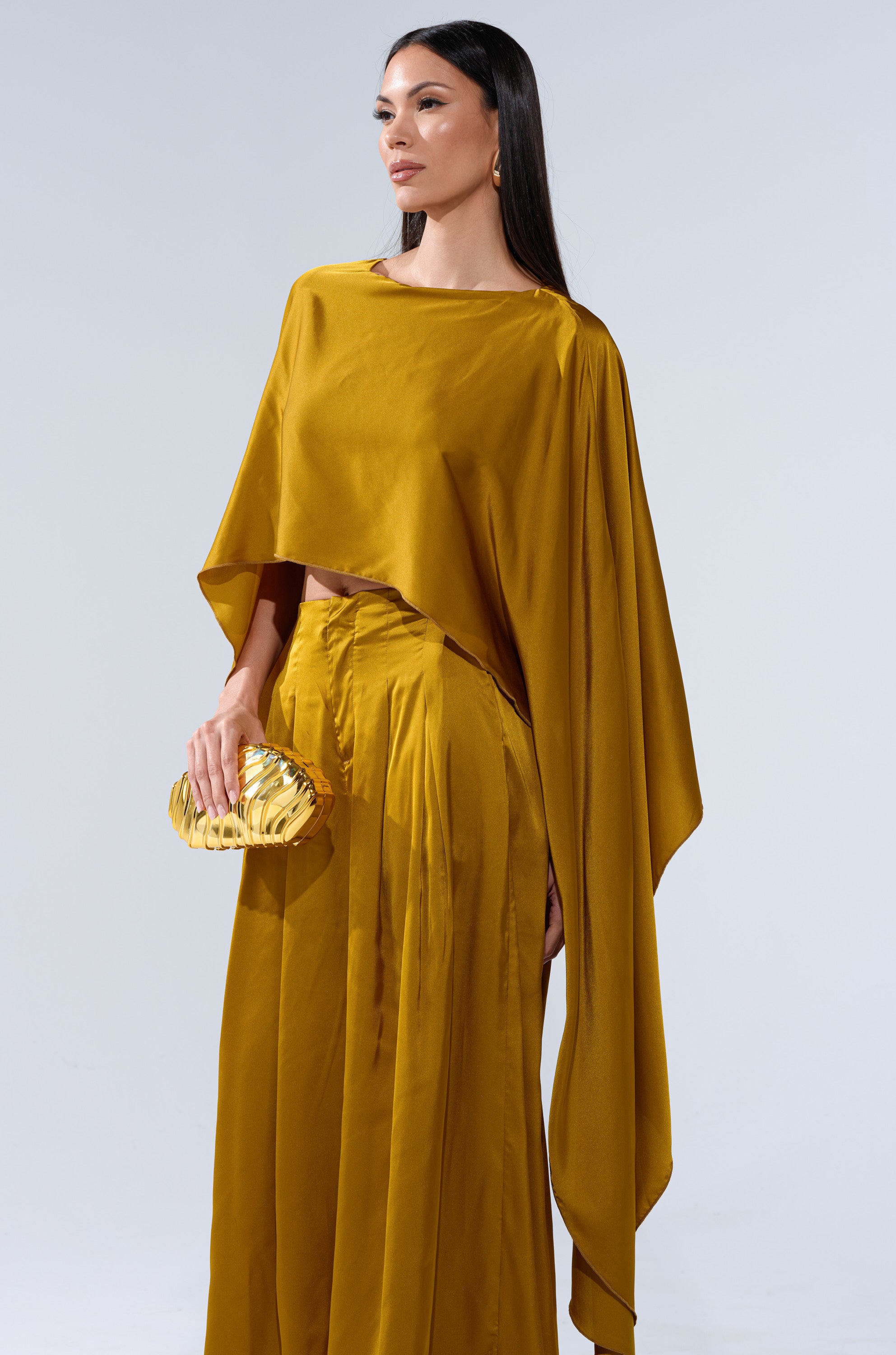 A woman with straight dark hair stands against a plain background, confidently wearing the COCKTAIL HOUR BLOUSE IN MUSTARD with wide-leg pants and holding a small gold clutch.