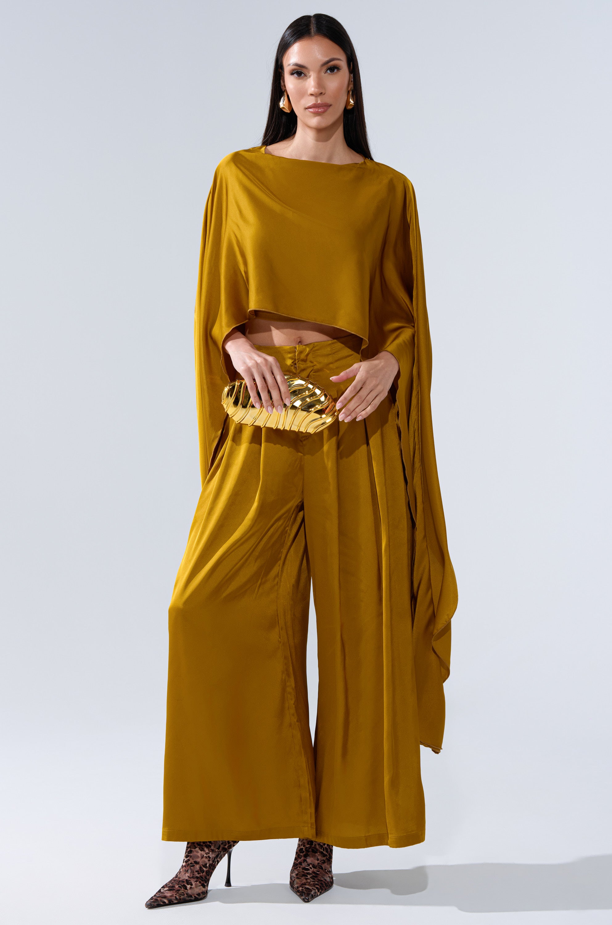 A woman stands against a plain backdrop wearing the COCKTAIL HOUR BLOUSE IN MUSTARD, paired with wide-leg pants. She accessorizes with a gold clutch, animal-print high heels, and hoop earrings.