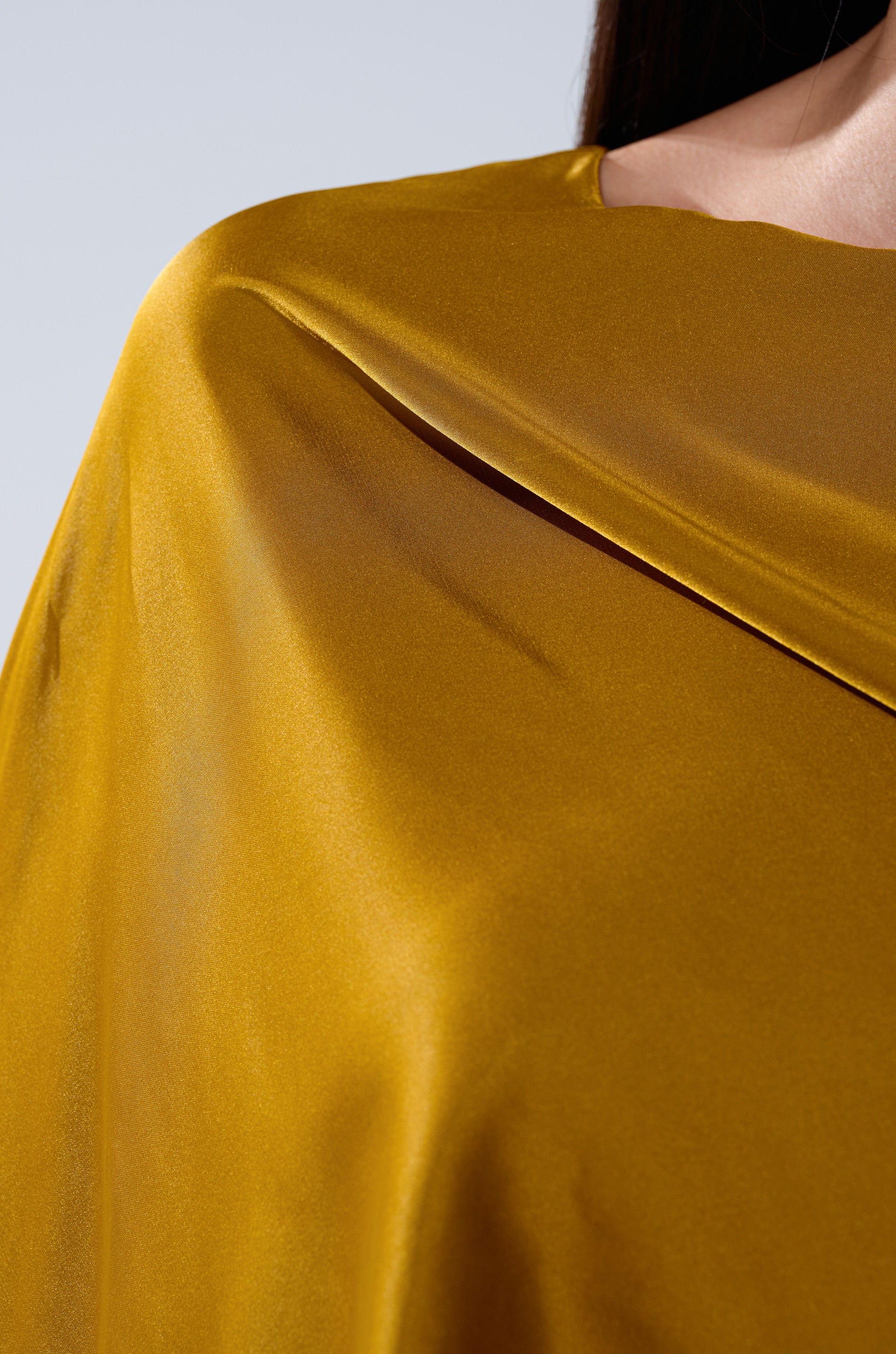 Close-up of a person wearing the COCKTAIL HOUR BLOUSE IN MUSTARD—a shiny, silky satin top with smooth folds and a soft sheen reflecting the light. The background is plain and neutral.