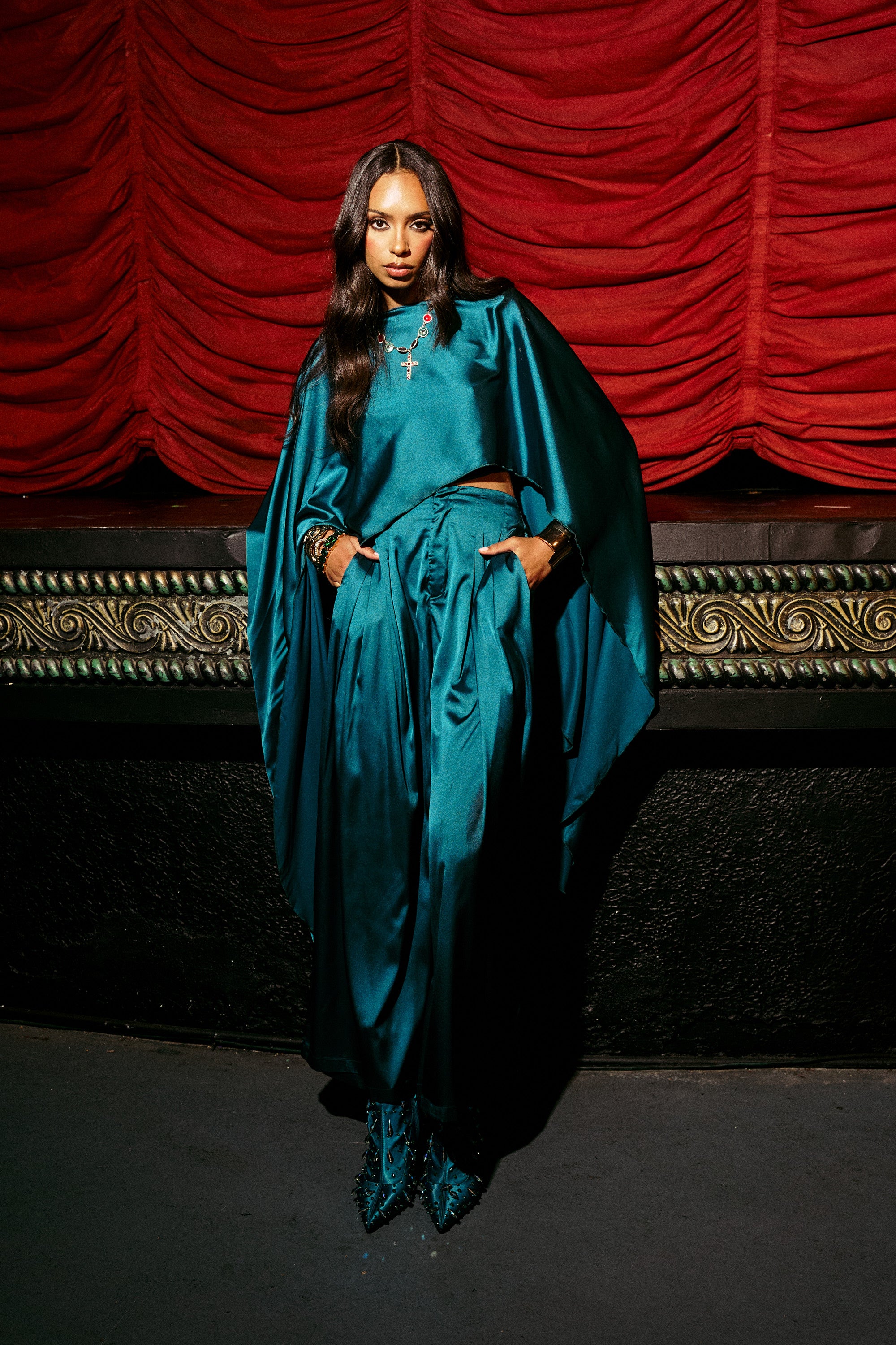 A woman with long dark hair and jewelry poses confidently with her hands in her pockets, wearing the GIRLS FOR LIFE EMERALD BLOUSE and high-waisted pants against a decorative stage with red velvet curtains.