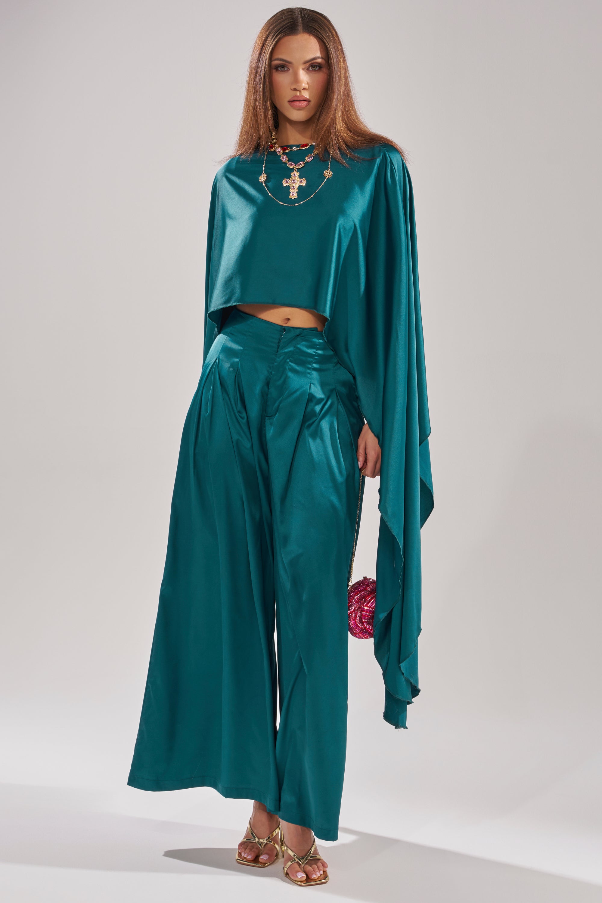 A woman stands facing the camera wearing the GIRLS FOR LIFE EMERALD BLOUSE, styled with wide-leg pants, a statement necklace, metallic heels, and a pink round clutch.