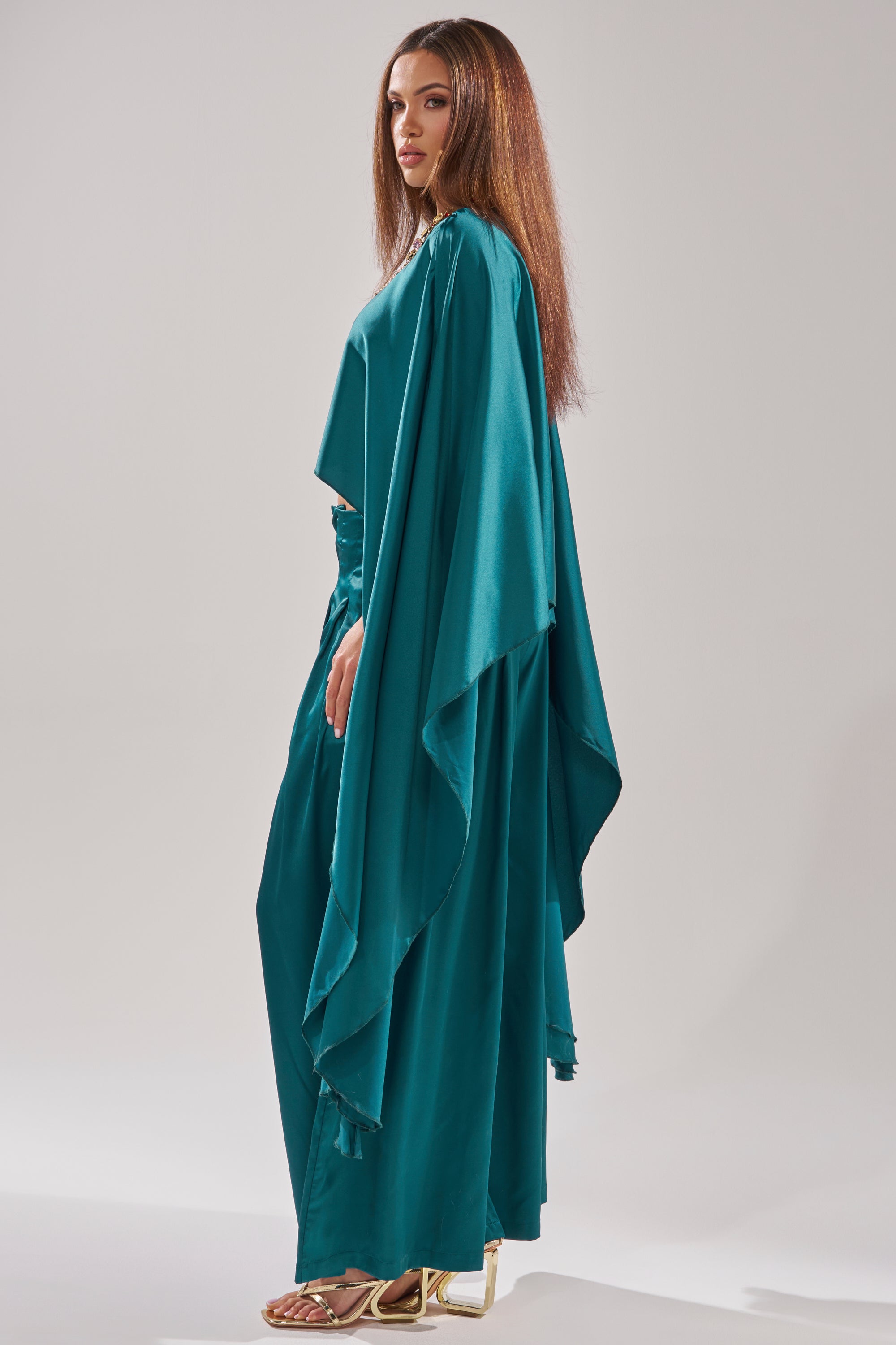A woman in profile wears the GIRLS FOR LIFE EMERALD BLOUSE, featuring draped sleeves and a high-low hem, paired with wide pants and gold heels. Her long brown hair is straight, set against a neutral background.
