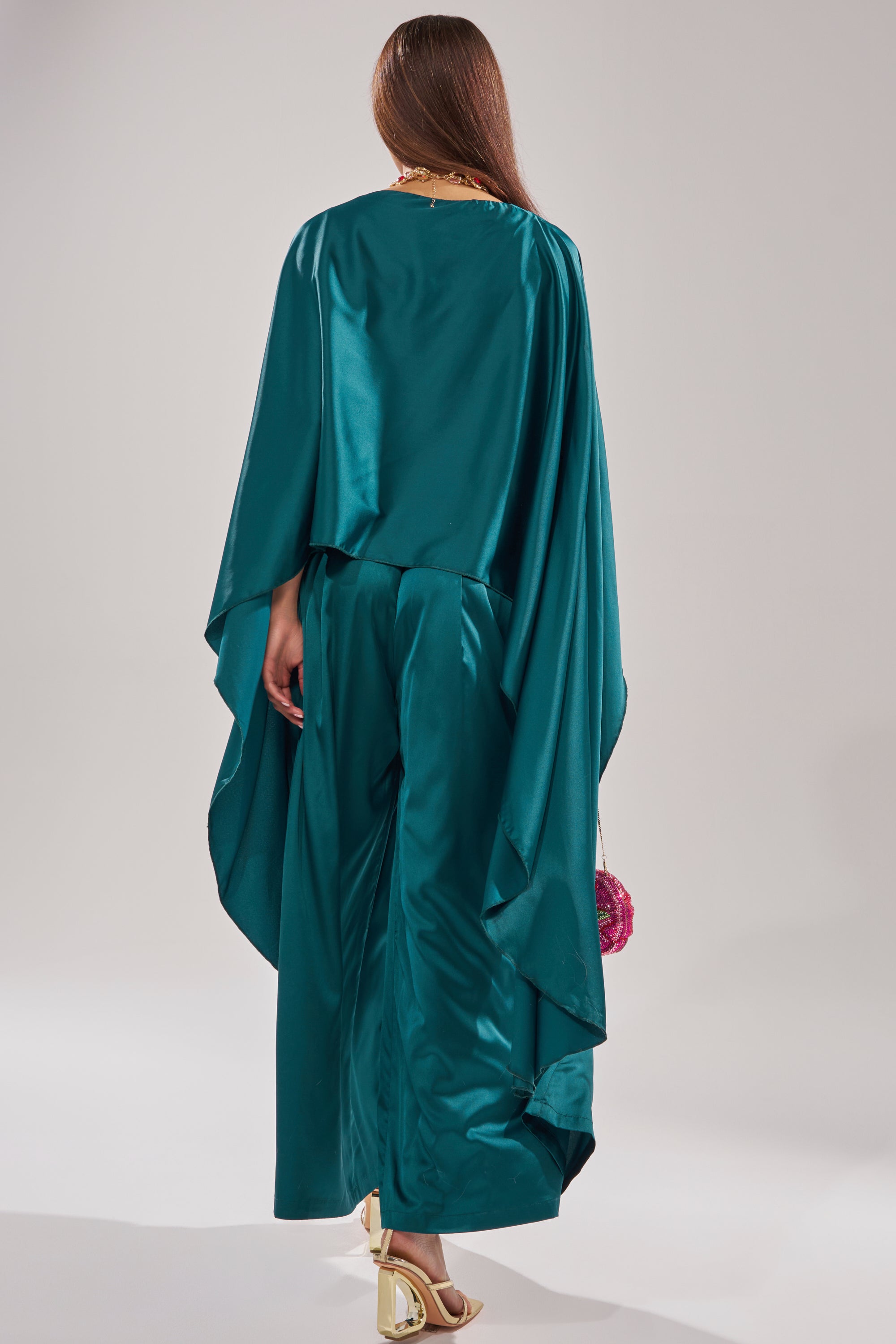 A woman with long brown hair faces away, wearing the GIRLS FOR LIFE EMERALD BLOUSE and matching wide-leg pants, holding a small pink bag and gold strappy heels, set against a plain light background.