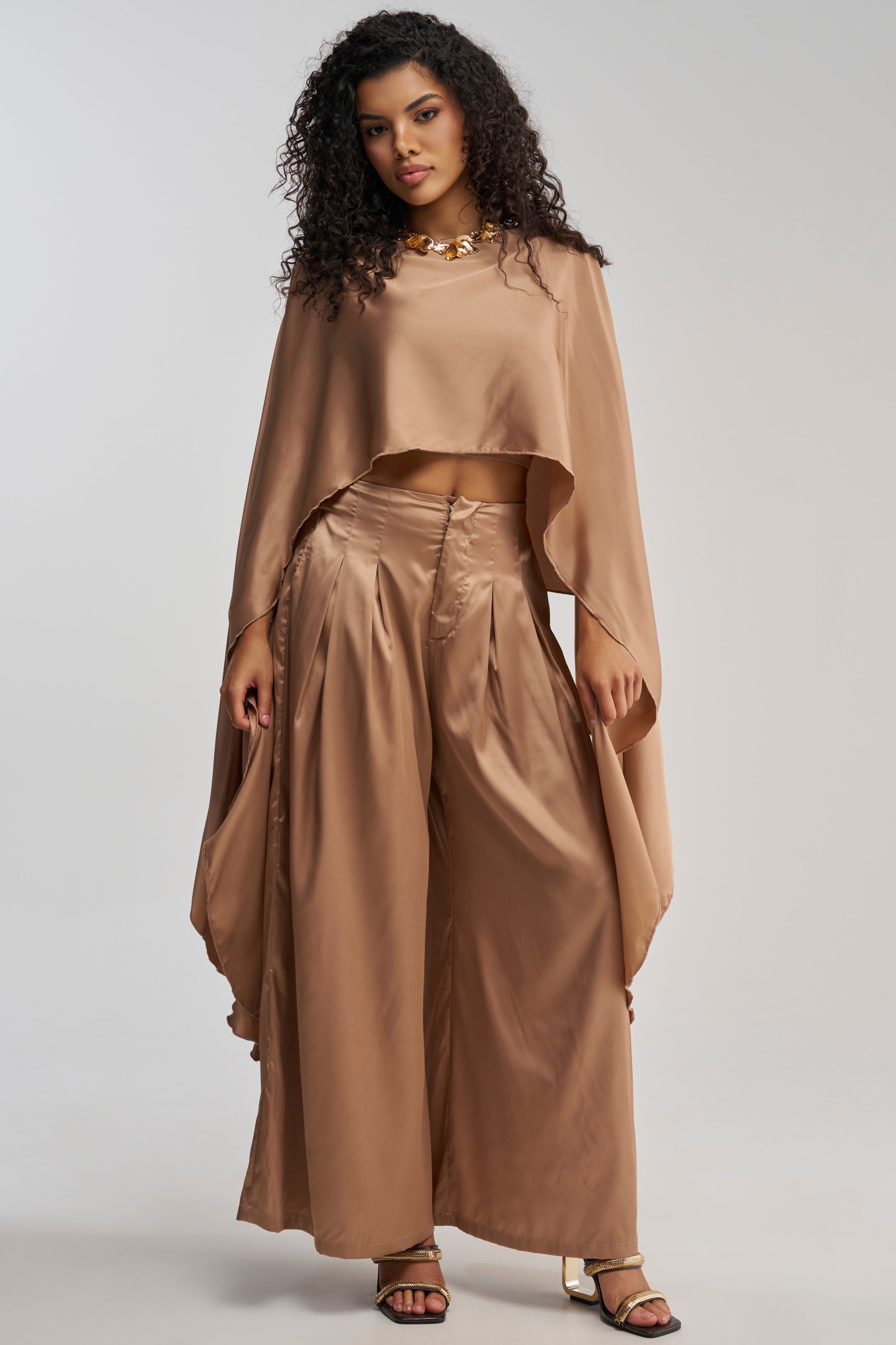 A woman with curly hair models the GOLDEN HOUR BLOUSE—a shiny, loose-fitting bronze top with a draped silhouette—paired with wide-leg pants, chunky necklace, and heeled sandals against a plain background.