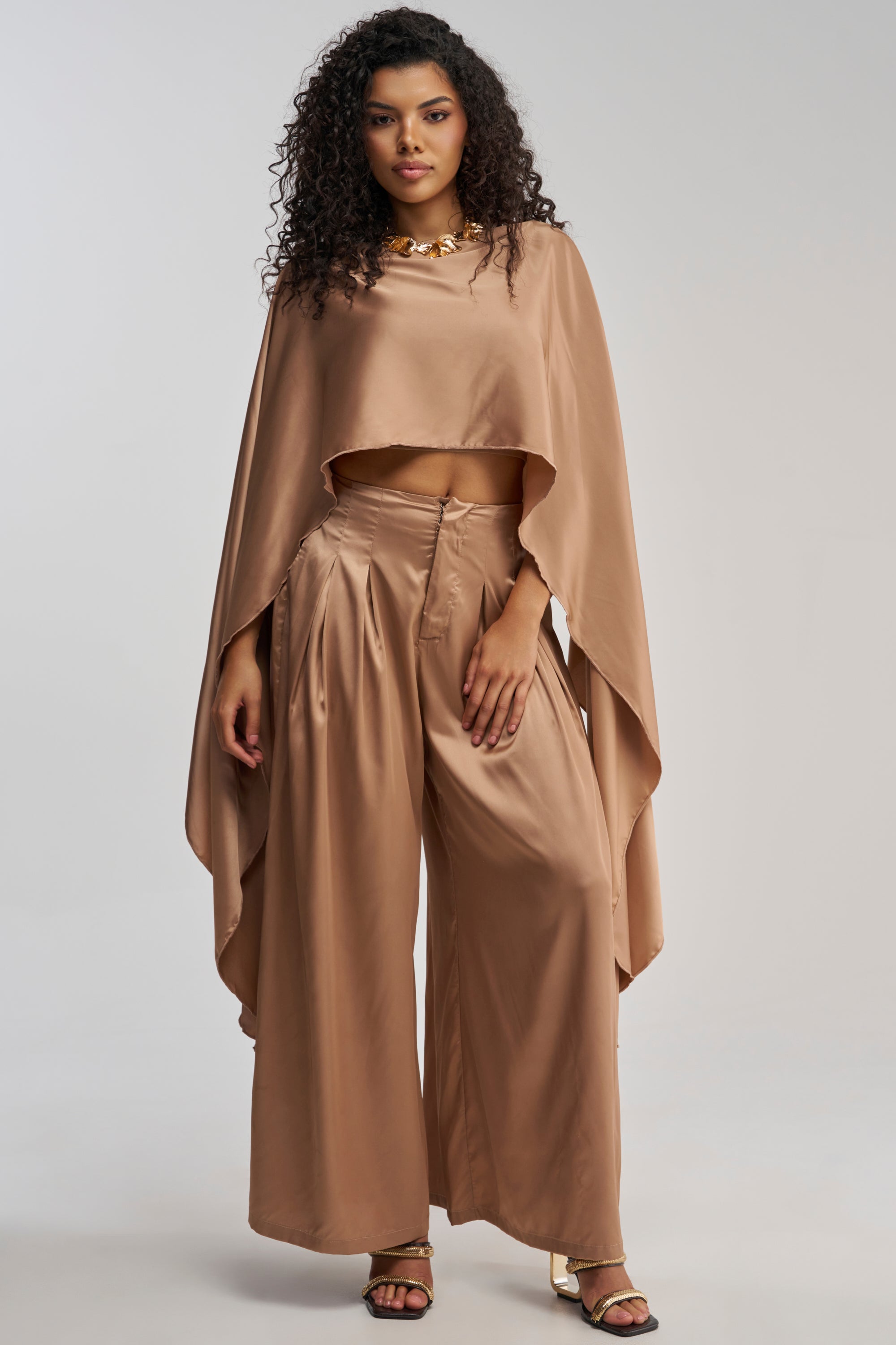 A woman stands confidently against a plain light background, her curly hair framing her face as she wears the GOLDEN HOUR BLOUSE—a shiny, light brown taupe satin top featuring wide, flowing handkerchief sleeves—paired with matching high-waisted, wide-leg pants and gold sandals.