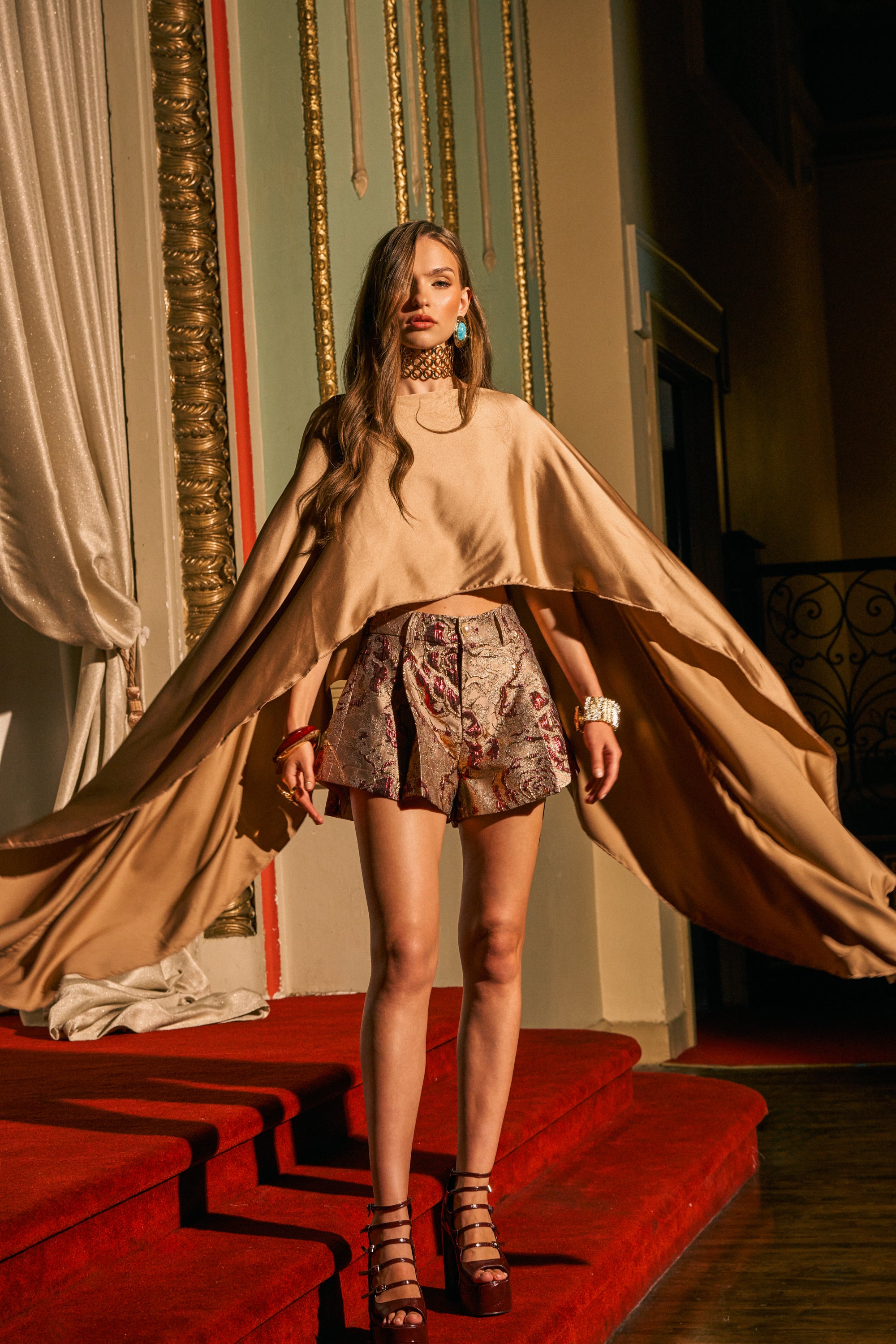 Wearing the GOLDEN HOUR BLOUSE, a woman stands on red-carpeted stairs, the flowing tan top with handkerchief sleeves and patterned shorts accentuated by gold-trimmed walls and dramatic lighting that showcases her fashion-forward style.