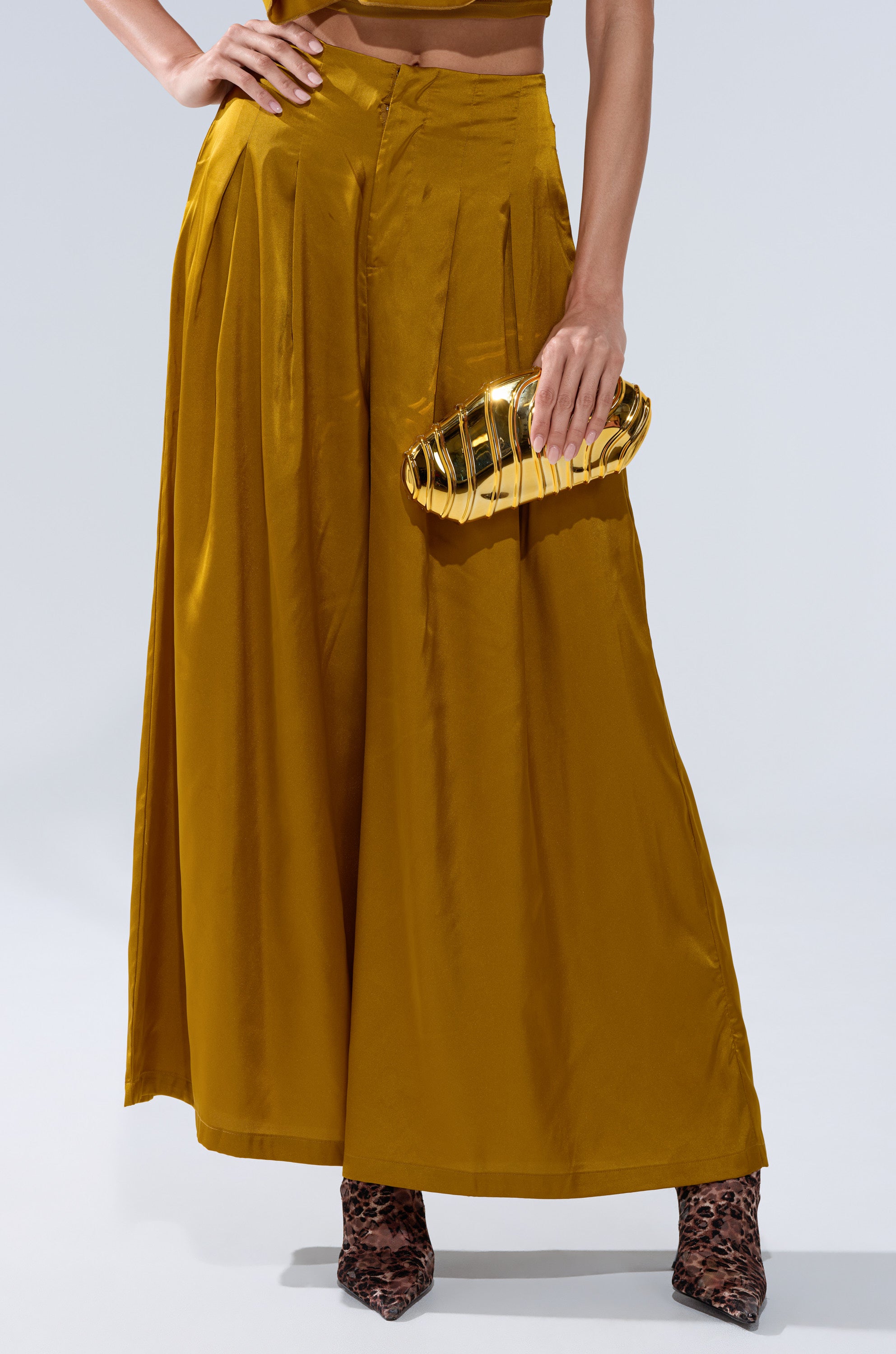 A person wears the COCKTAIL HOUR WIDE-LEG PANT IN MUSTARD with white nails, pointed-toe animal print shoes, and holds a shiny gold clutch. Their upper body is partially out of frame.