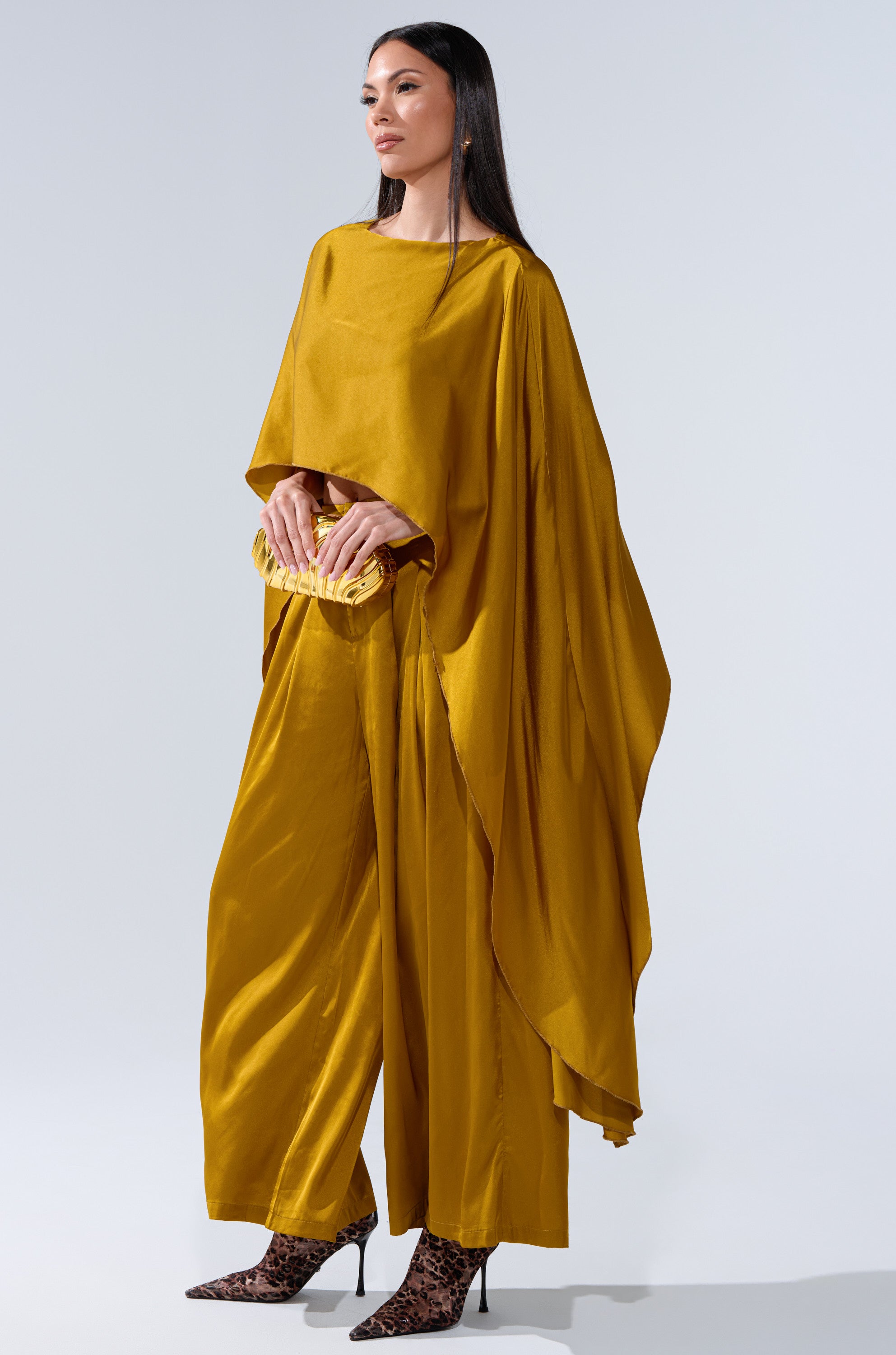 A woman models the COCKTAIL HOUR WIDE-LEG PANT IN MUSTARD with a matching draped top, pairing the high-rise, wide-leg pants with a small yellow clutch and leopard print high-heeled boots against a plain light background.