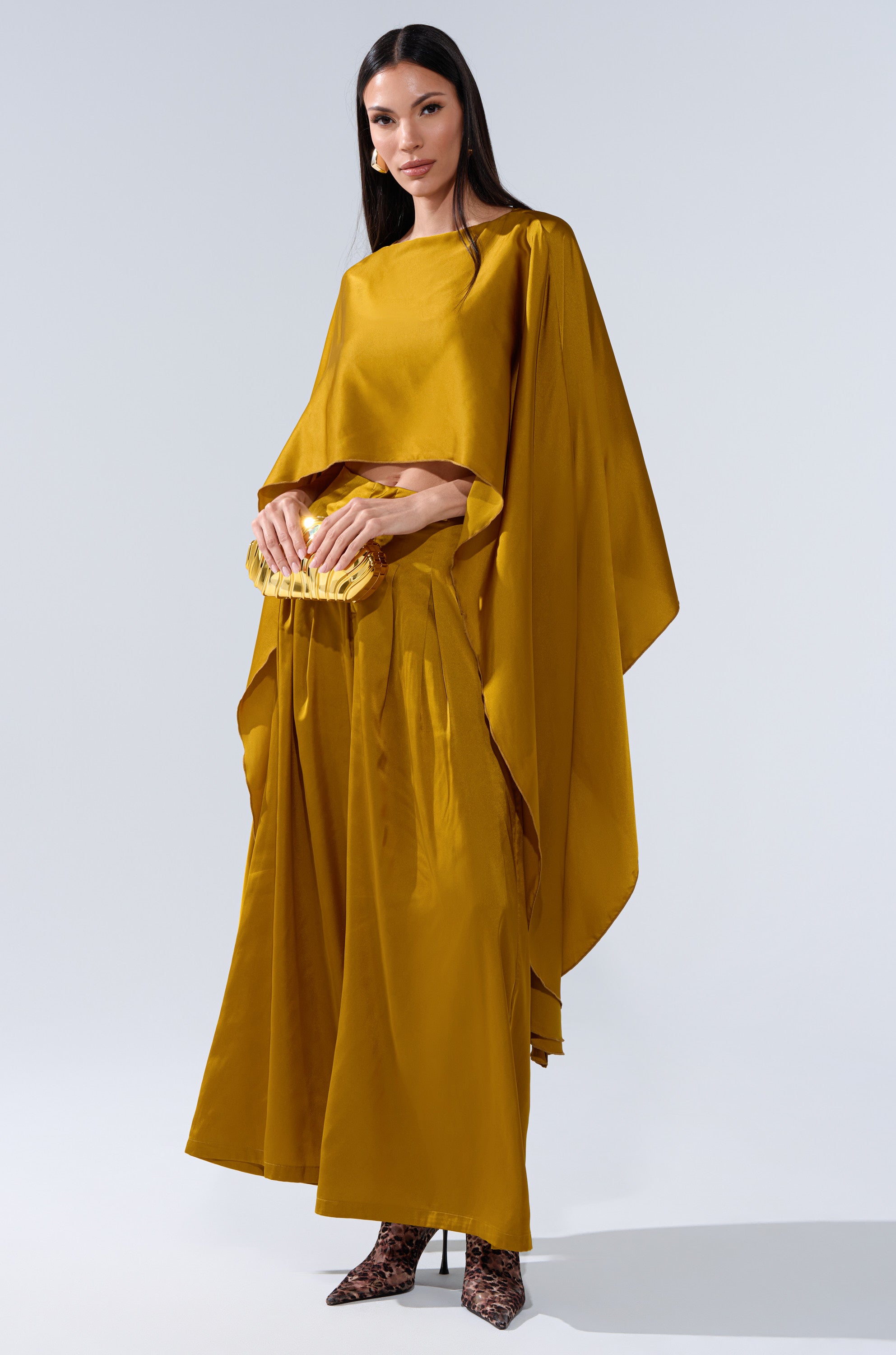 A woman poses against a plain backdrop in the COCKTAIL HOUR WIDE-LEG PANT IN MUSTARD, paired with a matching top. She accessorizes with a small yellow clutch and patterned high-heeled shoes.