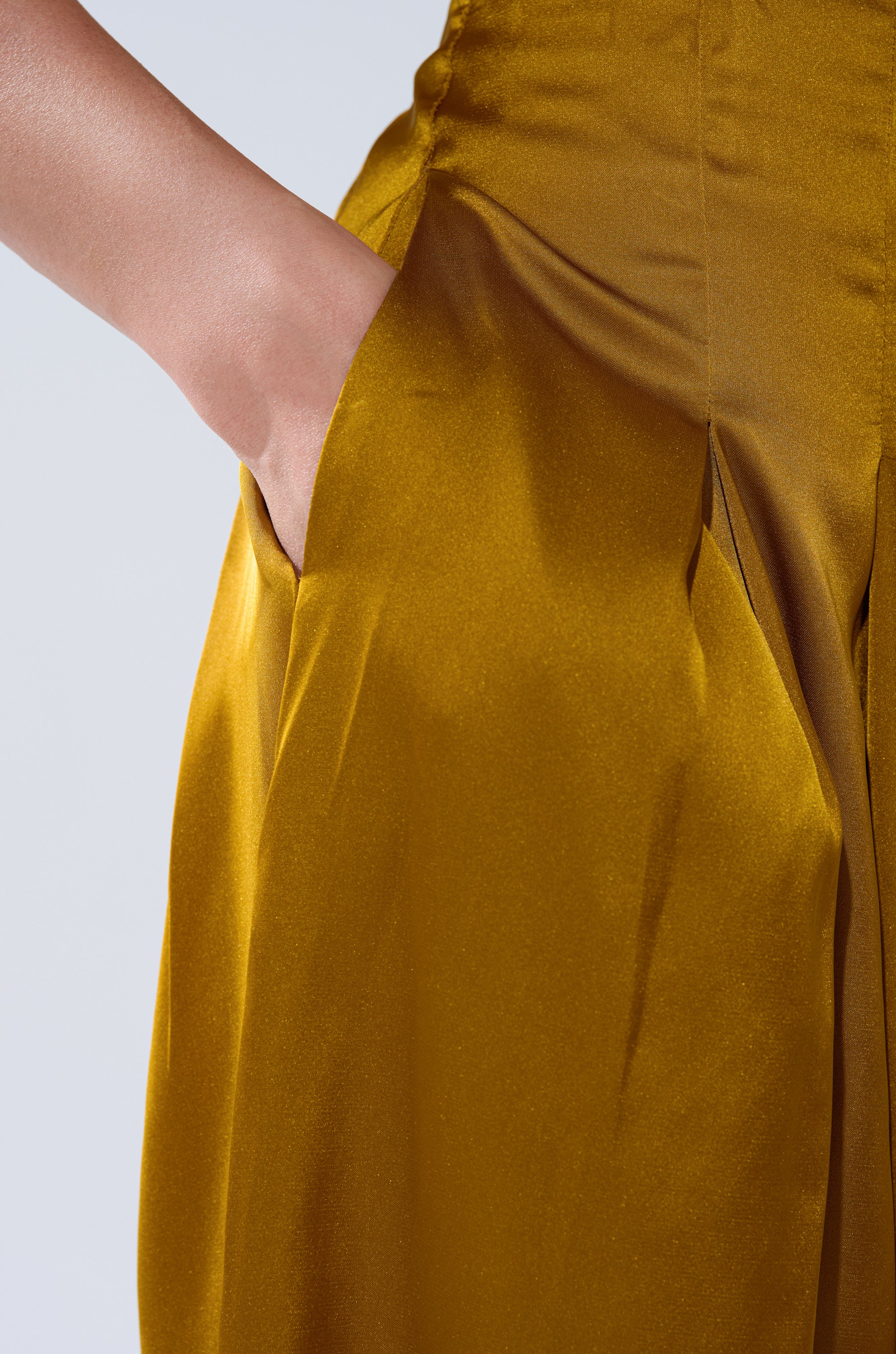 Wearing the COCKTAIL HOUR WIDE-LEG PANT IN MUSTARD, featuring a shiny golden satin finish and high-rise waistband, with one hand in the pocket, stands against a light background.
