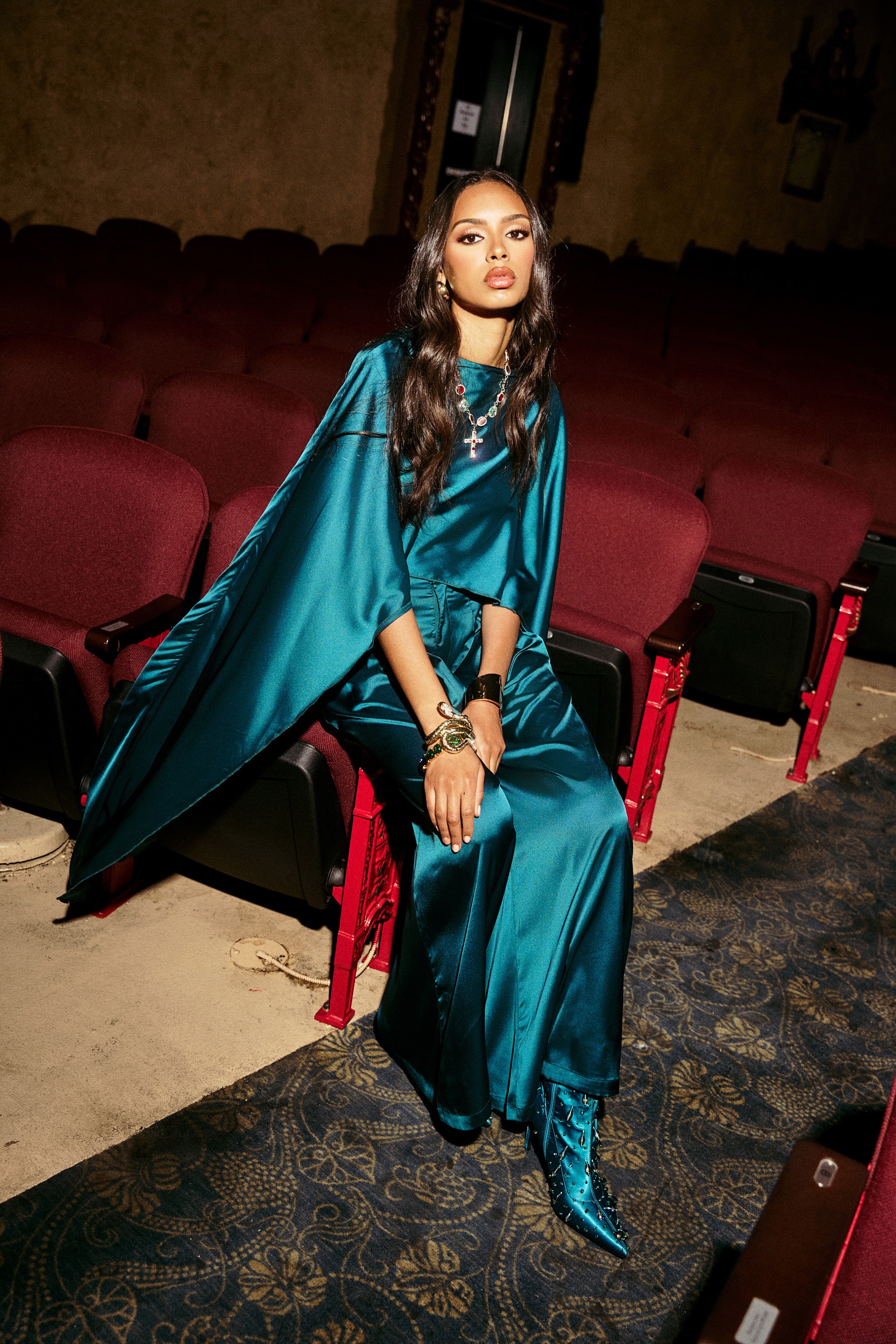 A woman wearing GIRLS FOR LIFE EMERALD TROUSER and shiny teal boots sits confidently on a theater chair in an empty auditorium with maroon seats and dim lighting.