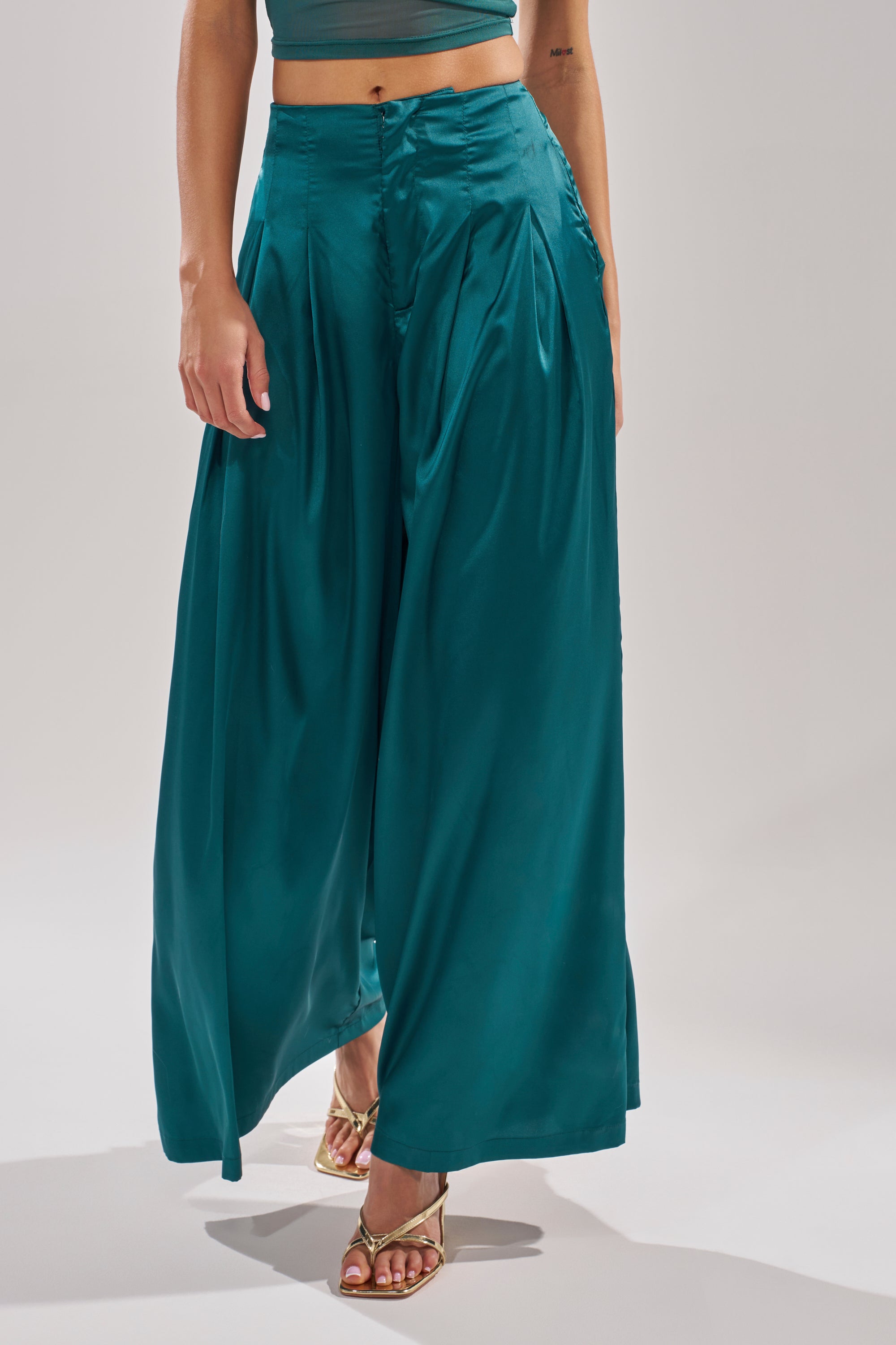 A person wears the GIRLS FOR LIFE EMERALD TROUSER—wide-leg satin pants in teal—with gold high-heeled sandals and a matching teal crop top. The photo is cropped from shoulders down against a plain light background. 1