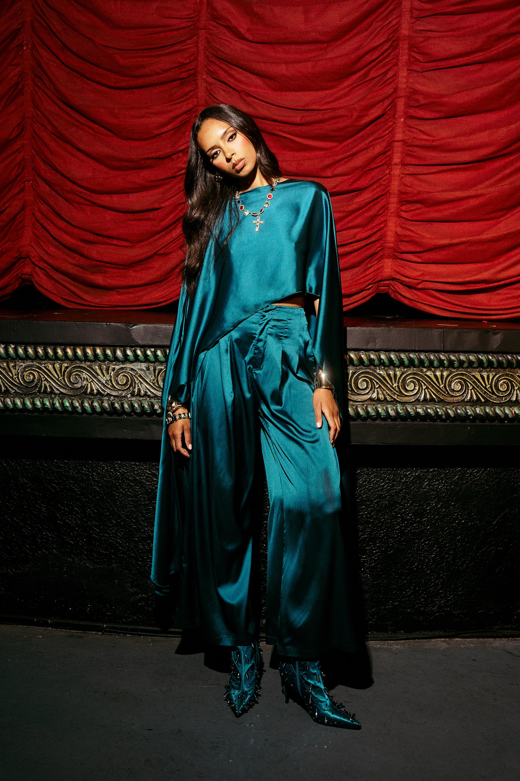 A woman confidently stands before a red theater curtain in the GIRLS FOR LIFE EMERALD TROUSER, featuring shiny teal satin fabric, a wide-leg silhouette, and matching boots, her long dark hair and jeweled accessories glowing in the stage light.