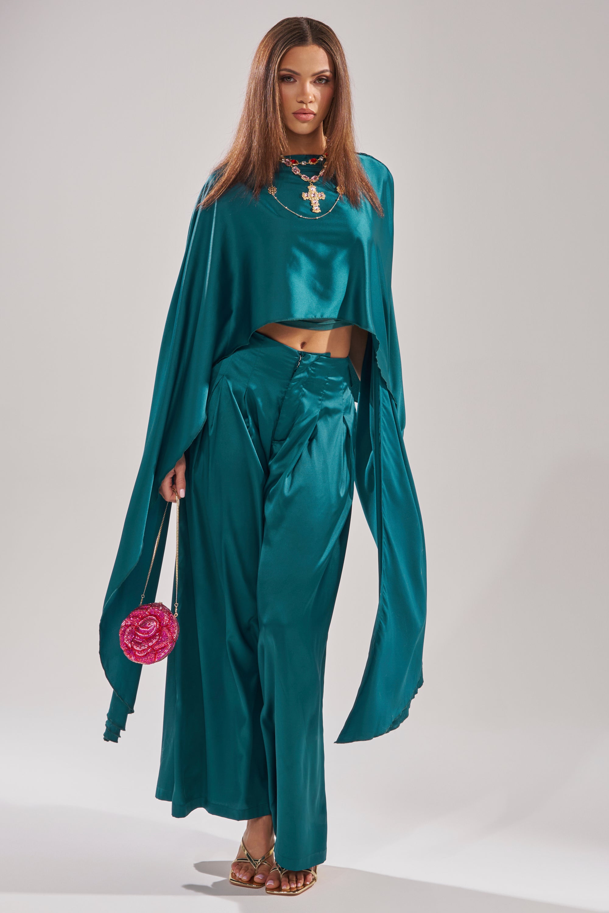 A woman poses against a plain light background in GIRLS FOR LIFE EMERALD TROUSER, paired with a teal satin cape top, statement necklace, gold sandals, and a pink rose-shaped handbag.