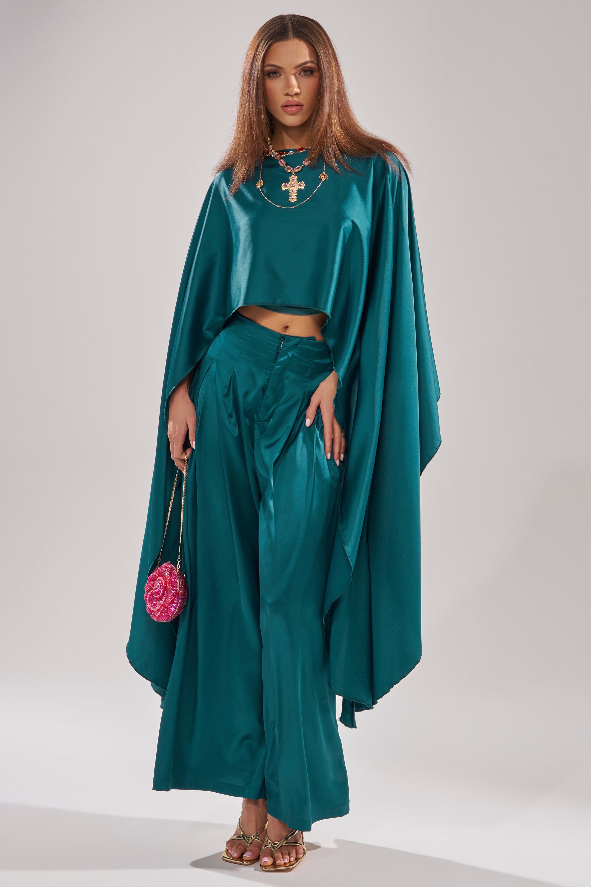 A woman stands confidently against a light background wearing the GIRLS FOR LIFE EMERALD TROUSER, a teal satin cape top, gold sandals, a statement necklace, and holding a small pink round purse.