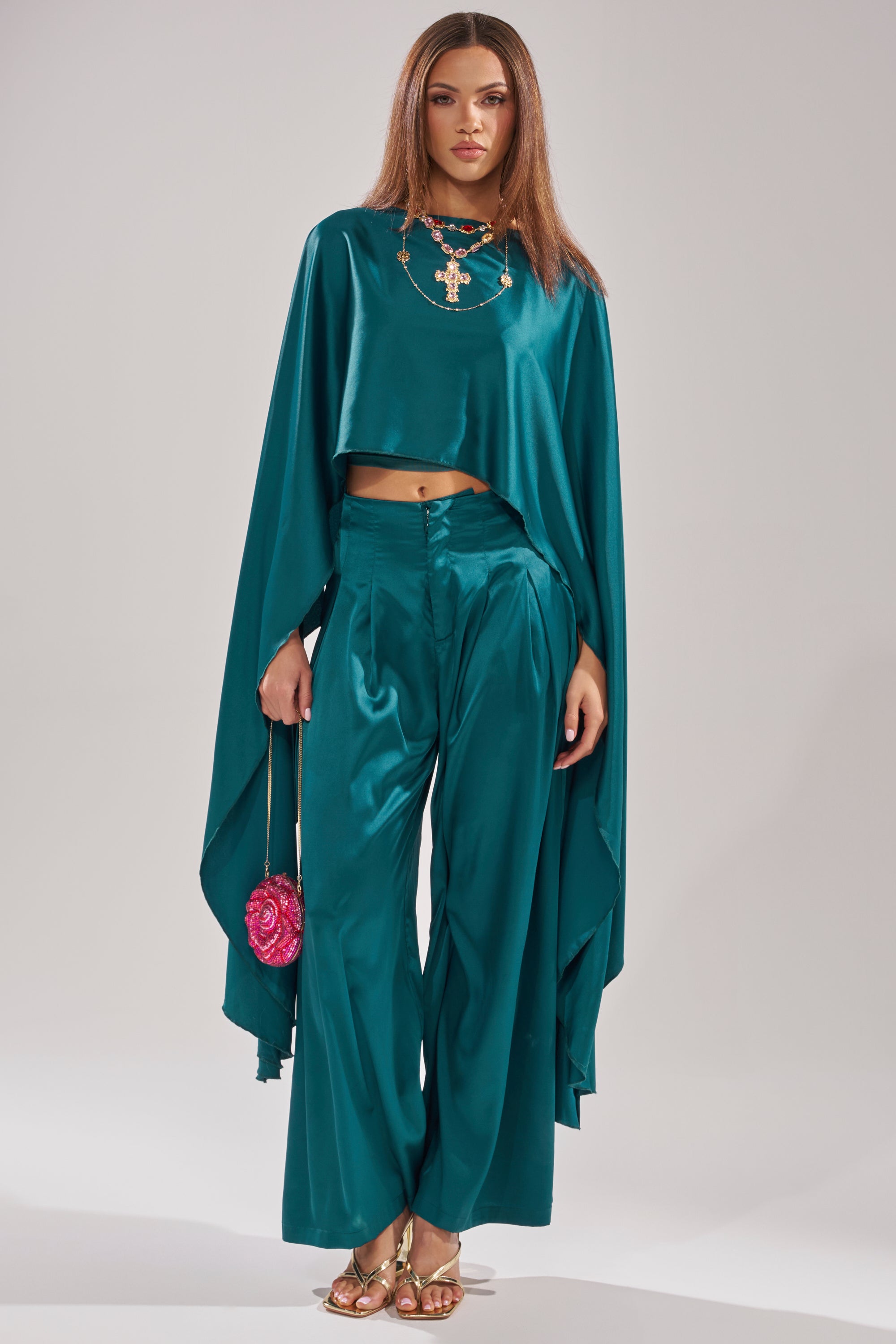 A woman wears the GIRLS FOR LIFE EMERALD TROUSER—a wide-leg satin pant—with a teal flowing cape top, gold sandals, statement necklace, and a small pink purse. Her straight hair is loose, and she has a neutral expression. 5