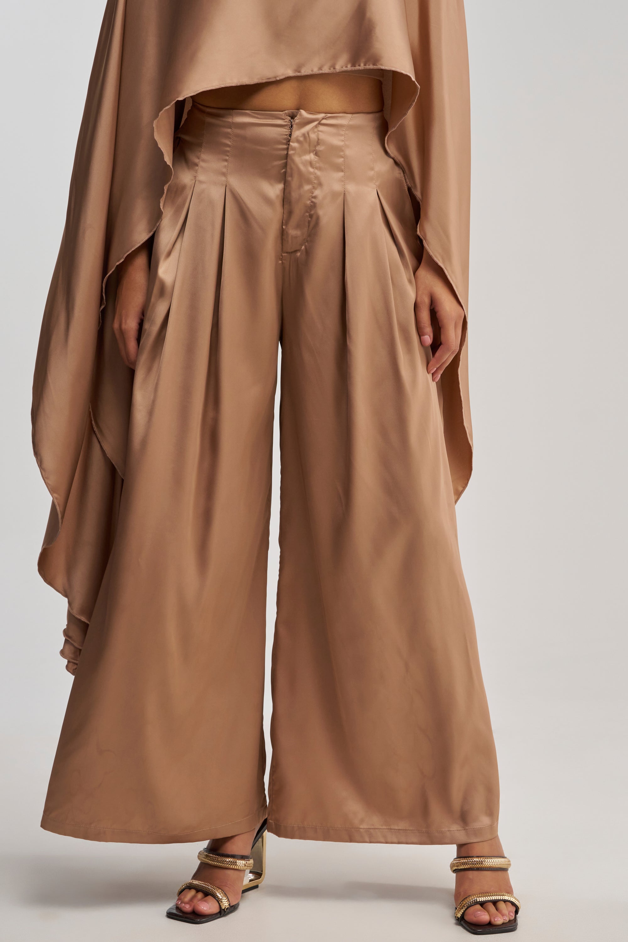 A person wearing the GOLDEN HOUR TROUSER, featuring a loose, high-rise fit in beige satin with a wide-leg silhouette and heeled sandals, complemented by a matching draped top partially visible.