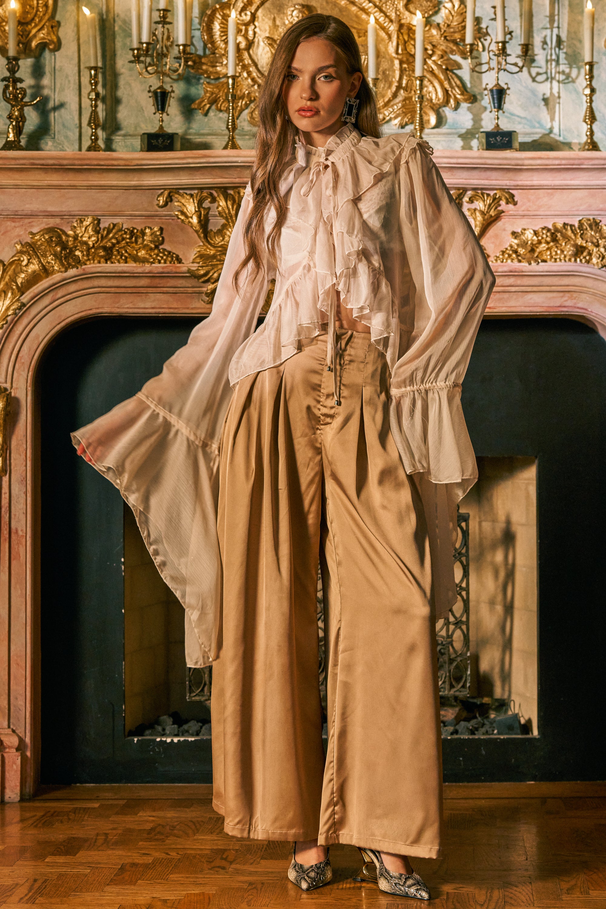 A woman stands in front of an ornate fireplace, wearing a sheer white ruffled blouse with wide sleeves and the GOLDEN HOUR TROUSER. Candles and gold accents decorate the mantel behind her.