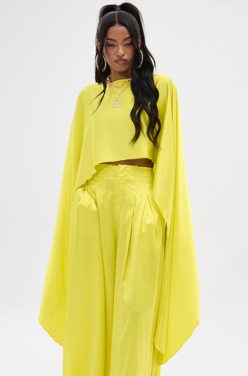 A person stands against a light background in a glossy satin outfit, featuring the COCKTAIL HOUR BLOUSE in bright yellow with flowing long sleeves and high-waisted, wide-leg pants, accessorized perfectly for cocktail hour with gold hoop earrings and layered necklaces.