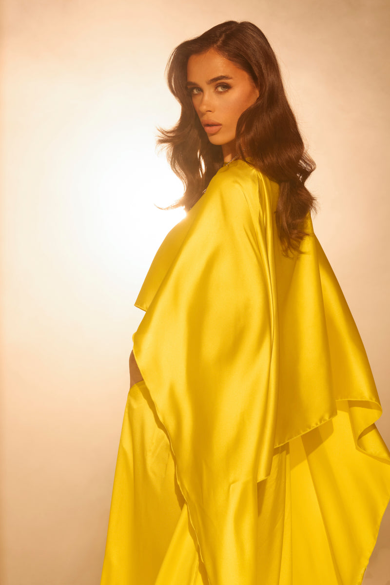 A woman with long, wavy brown hair poses in a glossy satin COCKTAIL HOUR BLOUSE by AKIRA Label against a warm, glowing background, softly illuminated from behind.