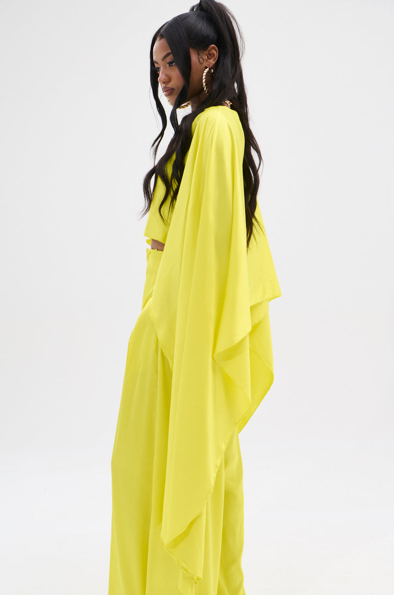 A woman with long dark hair poses in a vibrant yellow COCKTAIL HOUR BLOUSE featuring wide-leg pants and a flowing cape-like design with a high-low hem. Shining against the plain white background, she completes her look with hoop earrings and glossy satin fabric.