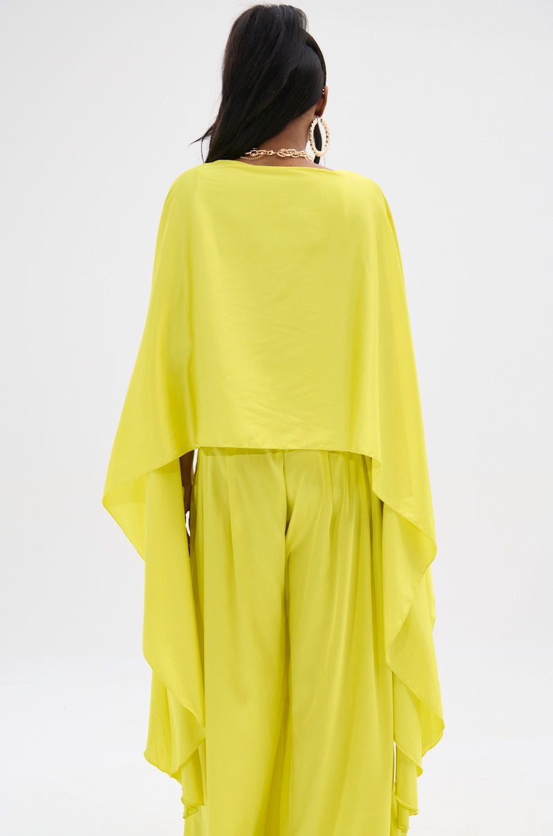 A person with long, dark hair faces away, wearing the COCKTAIL HOUR BLOUSE, a bright yellow, glossy satin cape-like top with a high-low hem, paired with matching wide-leg pants. Large hoop earrings and a chunky necklace complete the look against a plain white background.